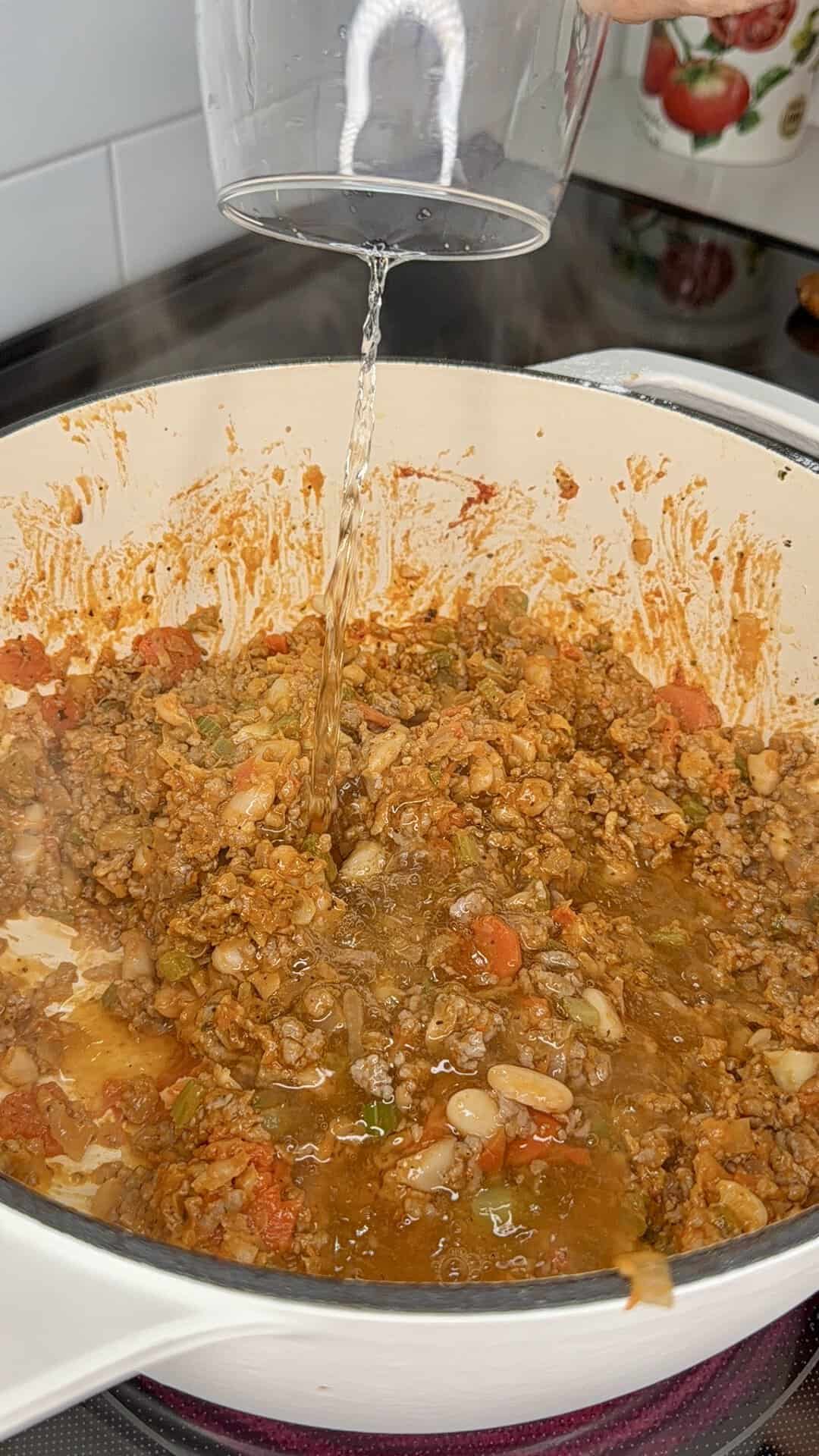 Water being poured from a clear glass into a Dutch oven filled with a mixture of ground meat, beans, and vegetables.