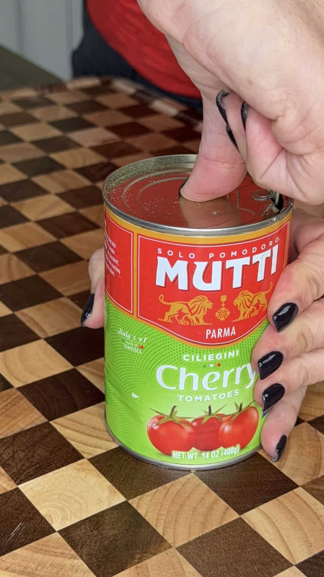 A hand opening a 14-ounce can of Mutti cherry tomatoes on a checkered wooden surface.