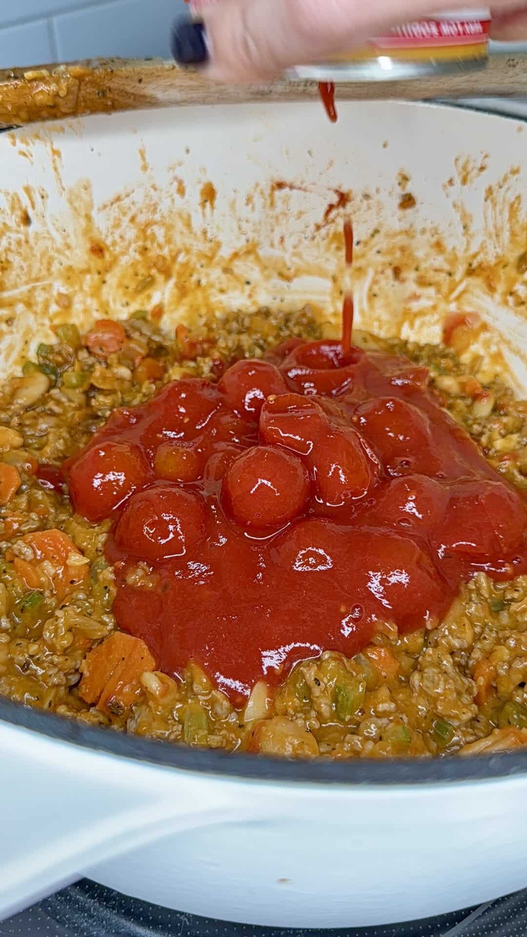 Canned cherry tomatoes being poured into a pot containing a mixture of ground meat and vegetables.