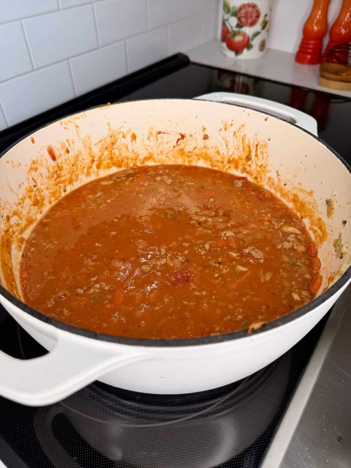 A white Dutch oven filled with a red meat-based pasta sauce sitting on a stovetop.