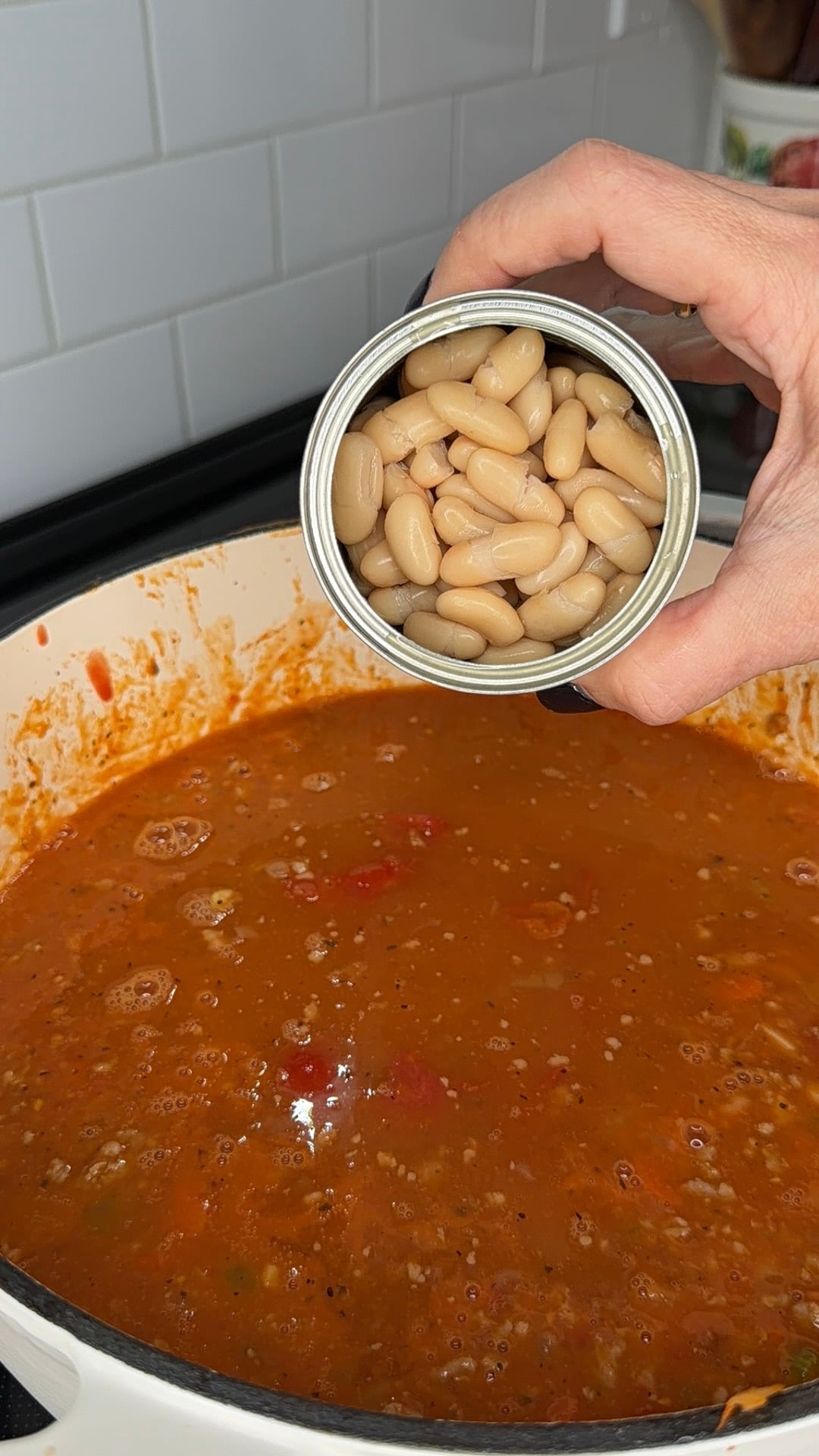 A hand holds an open can of white beans over a pot of simmering tomato-based stew.