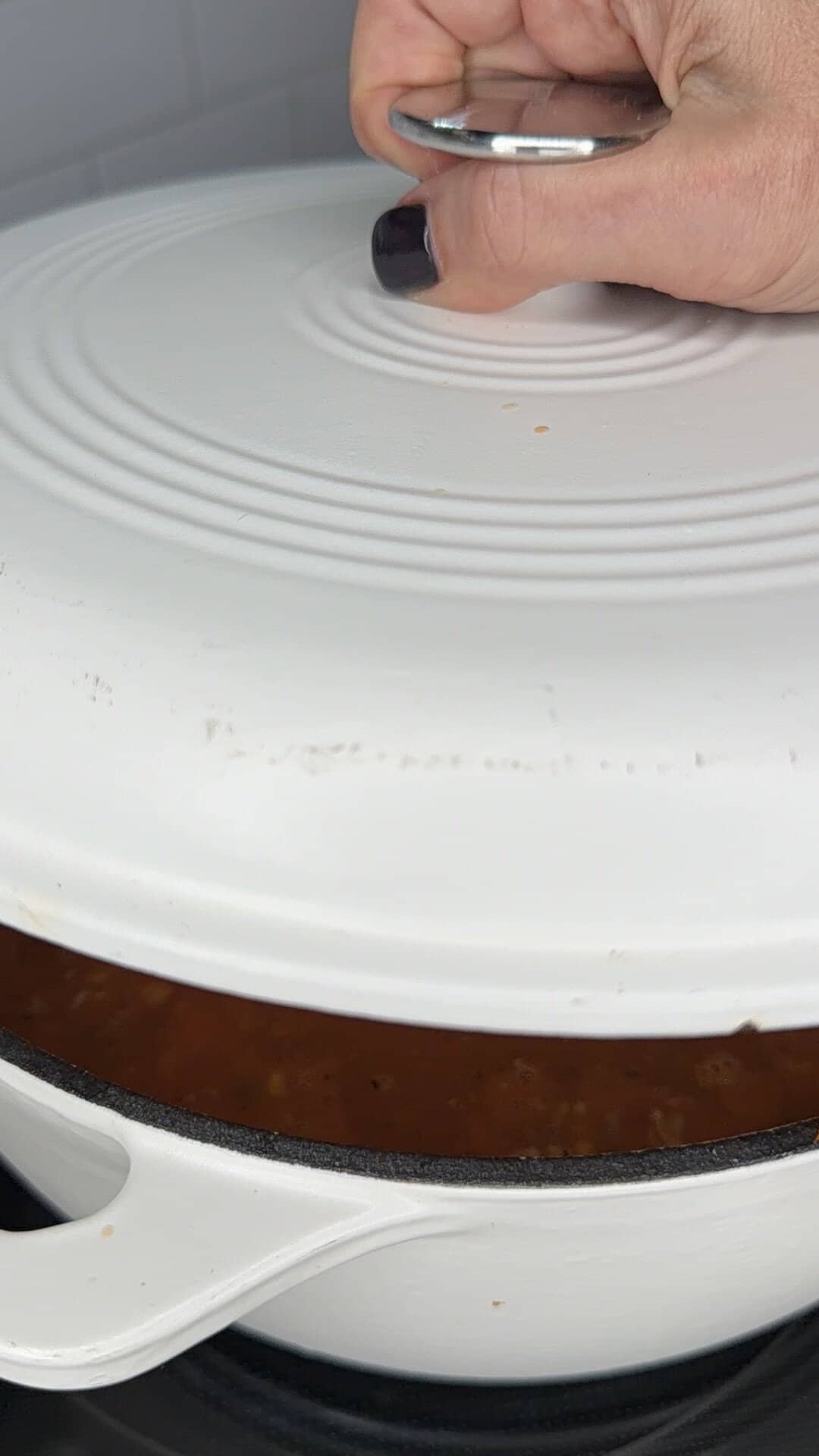 A hand lifts the white lid of a pot containing a dark, textured stew.