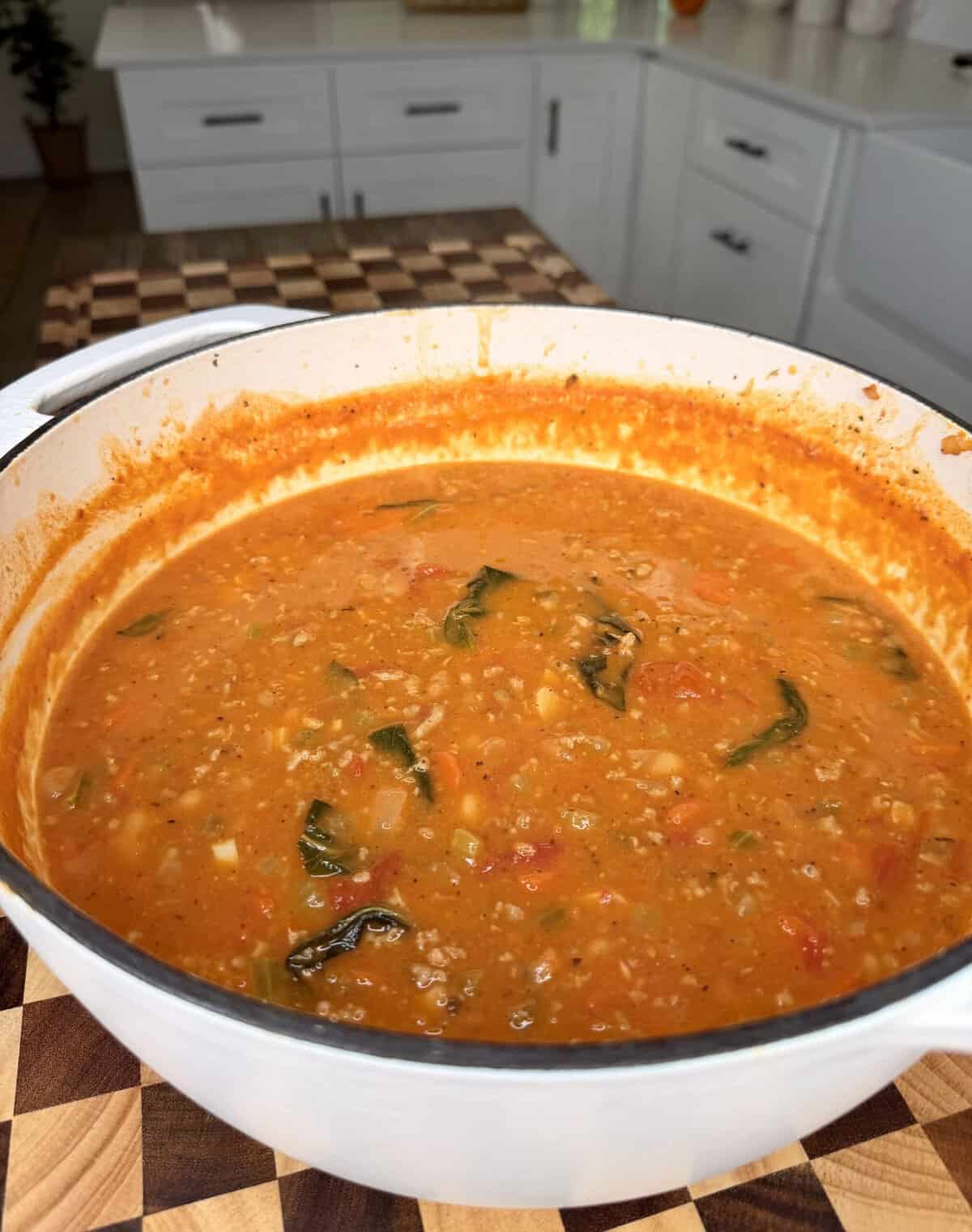 A white Dutch oven filled with tomato-based vegetable and grain soup containing spinach, resting on a wooden cutting board in a kitchen.