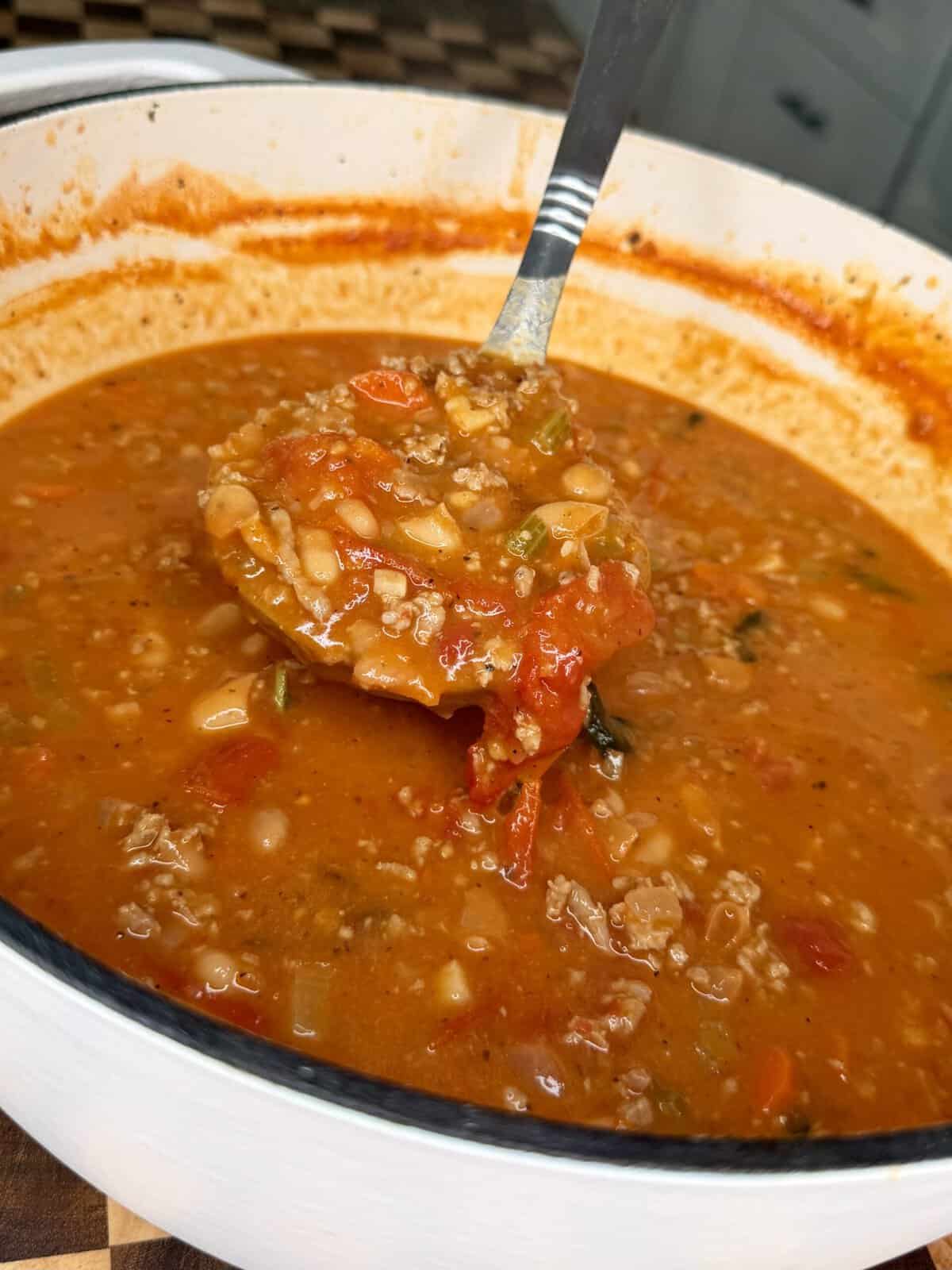 A metal ladle lifts a scoop of hearty vegetable and ground meat soup from a large white pot.