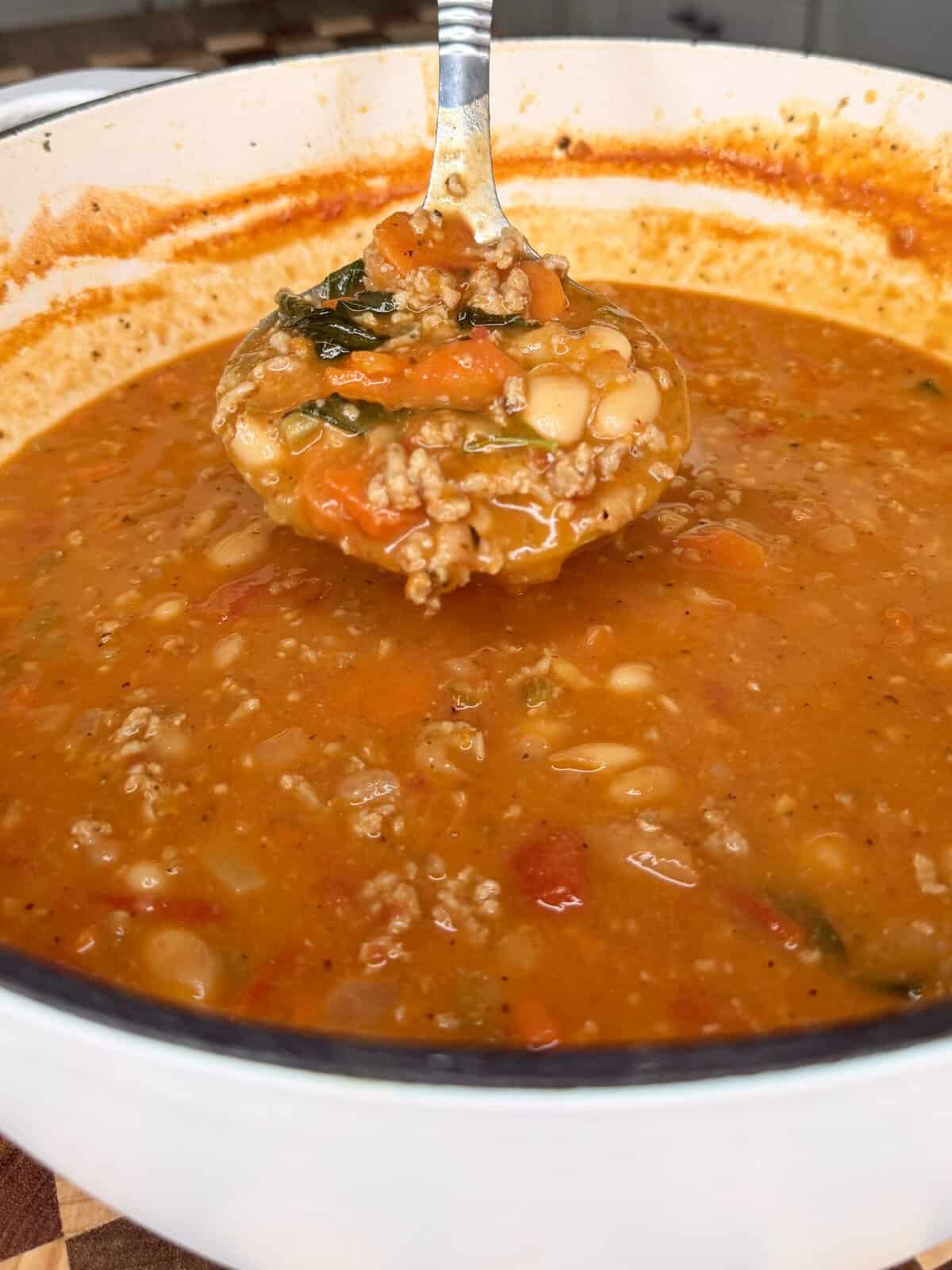 A metal ladle lifting a spoonful of hearty soup with ground meat, white beans, and vegetables from a white Dutch oven.