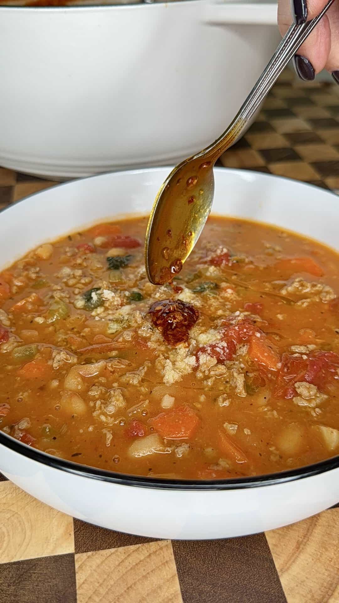A spoon drizzling chili oil into a bowl of sausage, white bean, and tomato soup.
