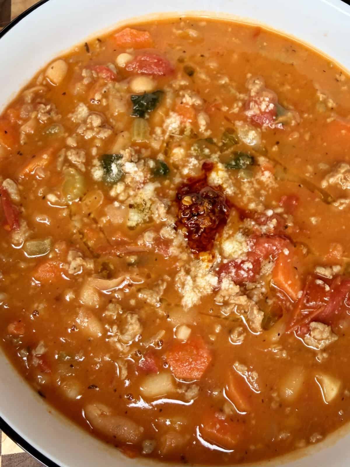 A bowl of vegetable and ground meat soup topped with grated cheese and a dollop of chili oil.