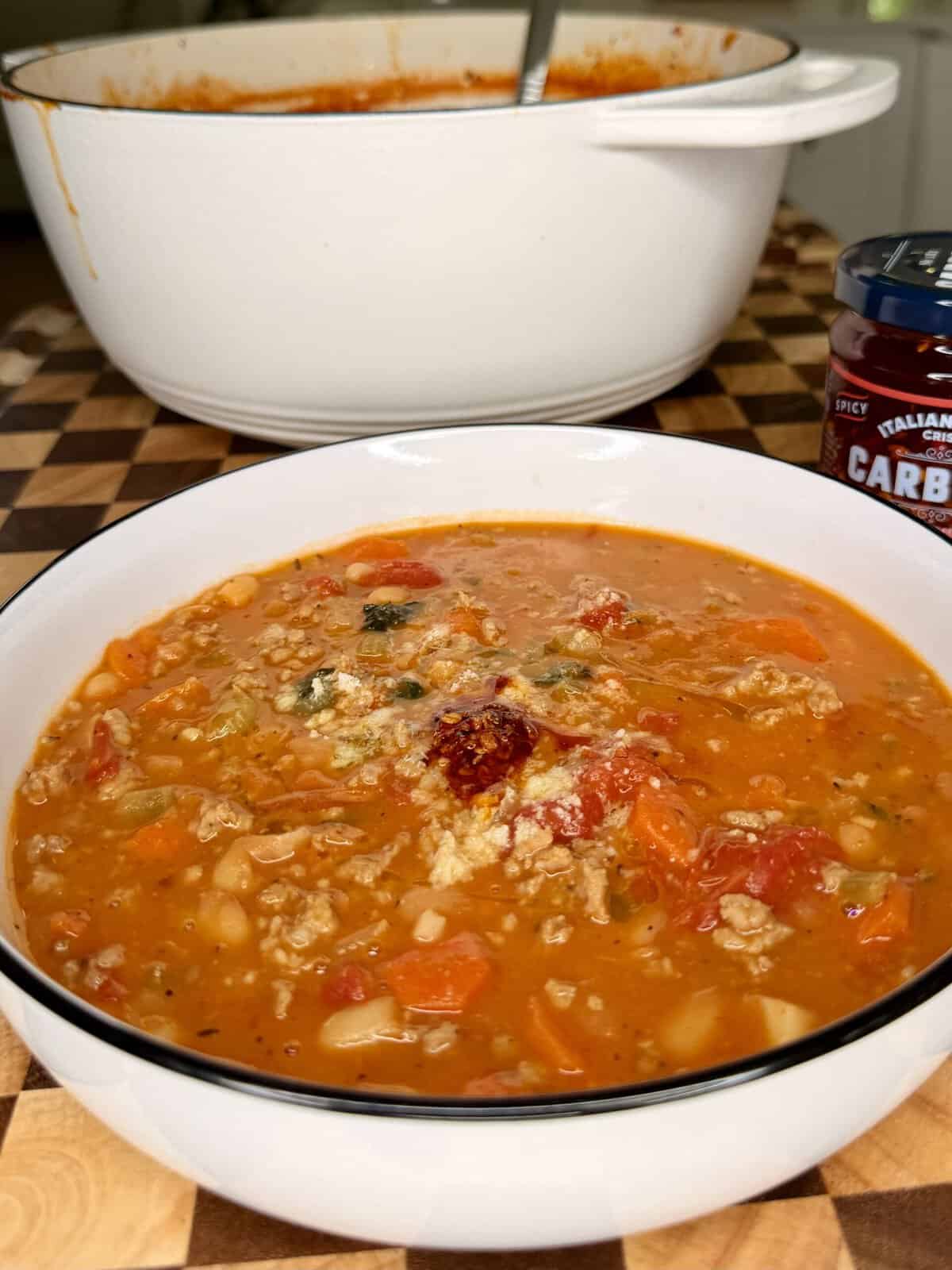 A bowl of chunky vegetable and ground meat soup with a spoonful of chili oil and grated cheese on top, with a large pot and a jar of chili crisps in the background.