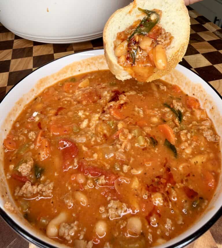 A slice of bread topped with bean and vegetable soup held over a bowl of the same hearty tomato-based soup.