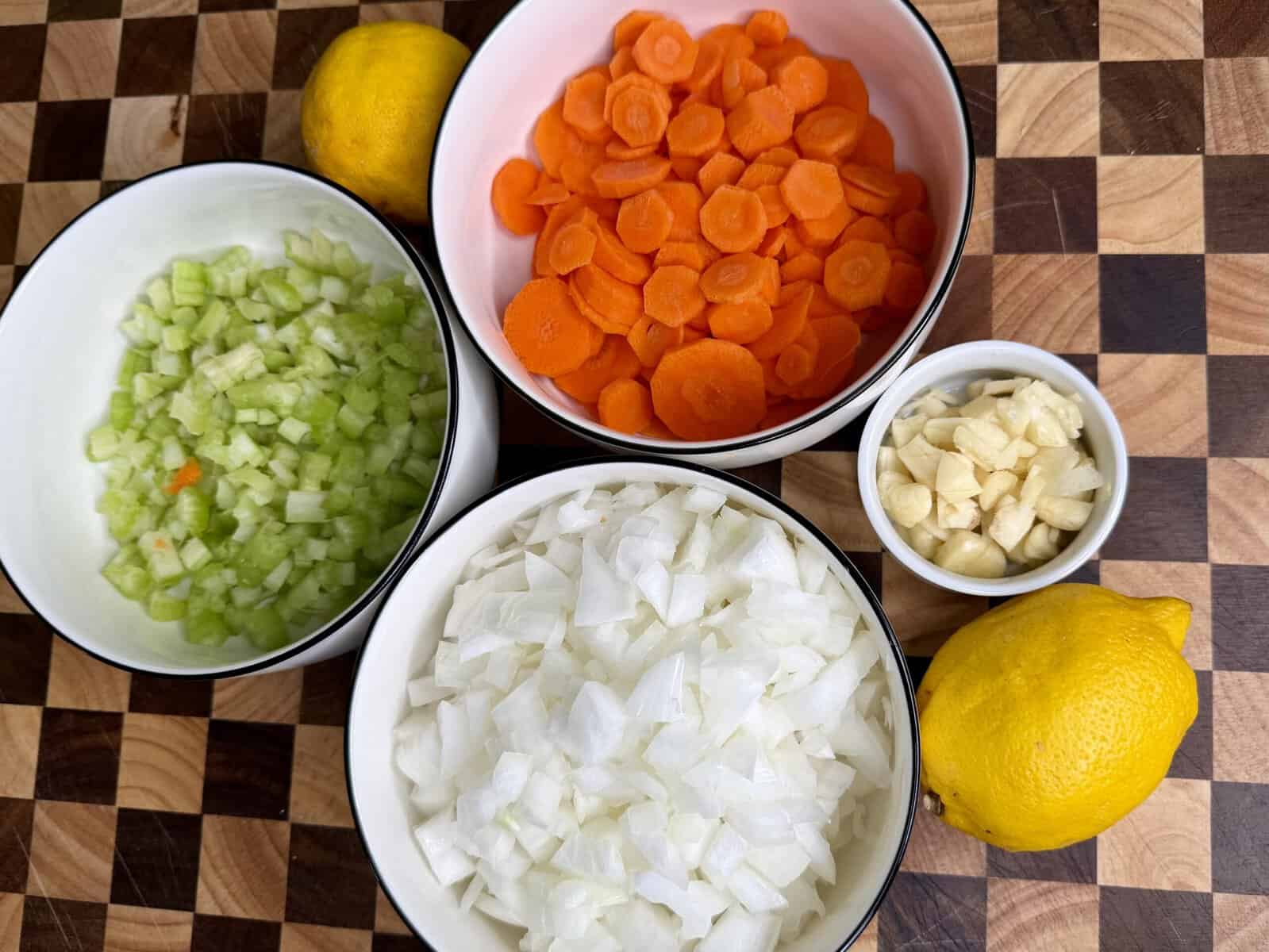 Bowls of chopped celery, sliced carrots, diced onions, and garlic cloves, with two whole lemons on a checkered cutting board.