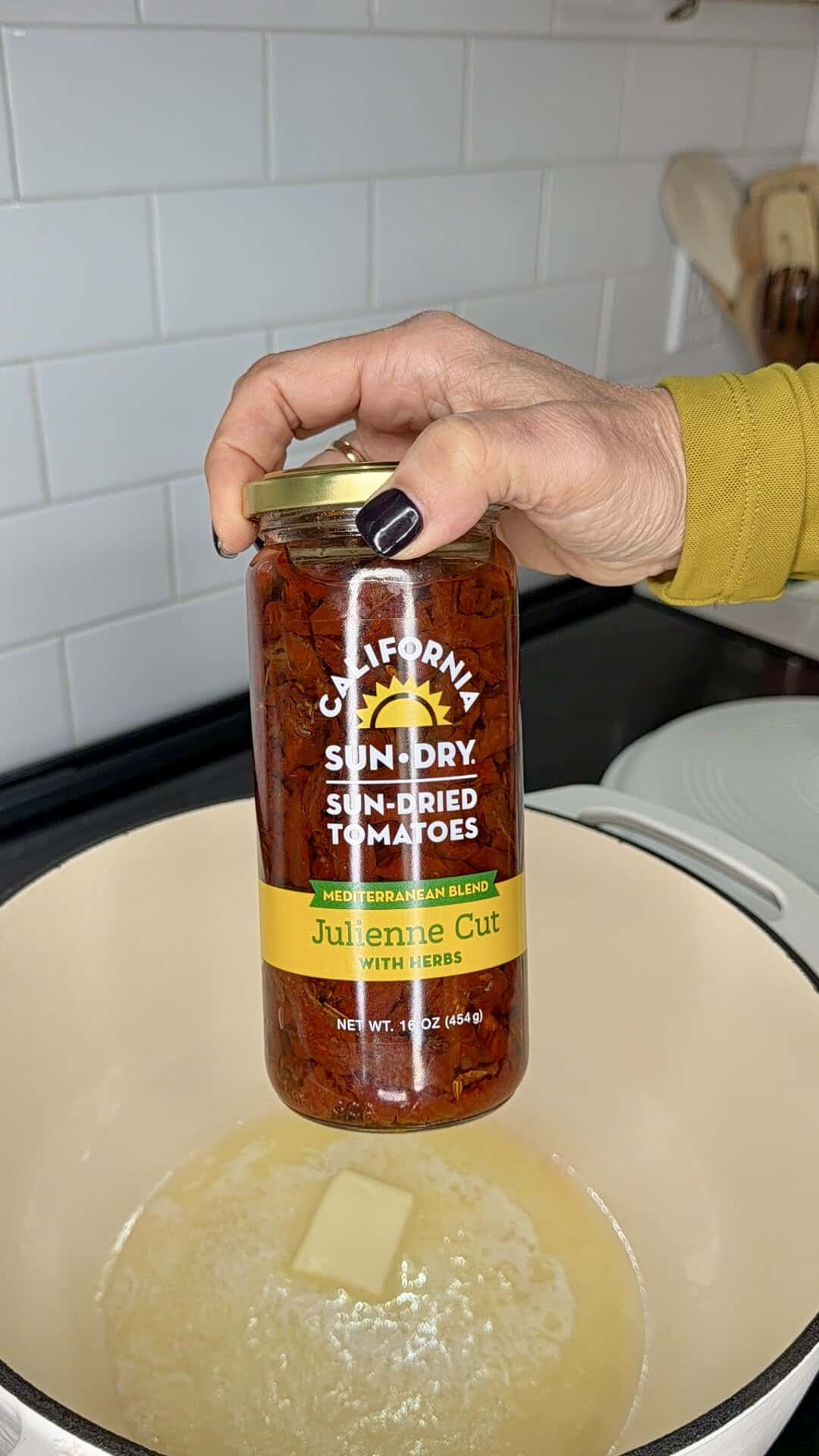 A hand holds a jar of California Sun-Dry Mediterranean blend julienne cut sun-dried tomatoes over a pot of melting butter.