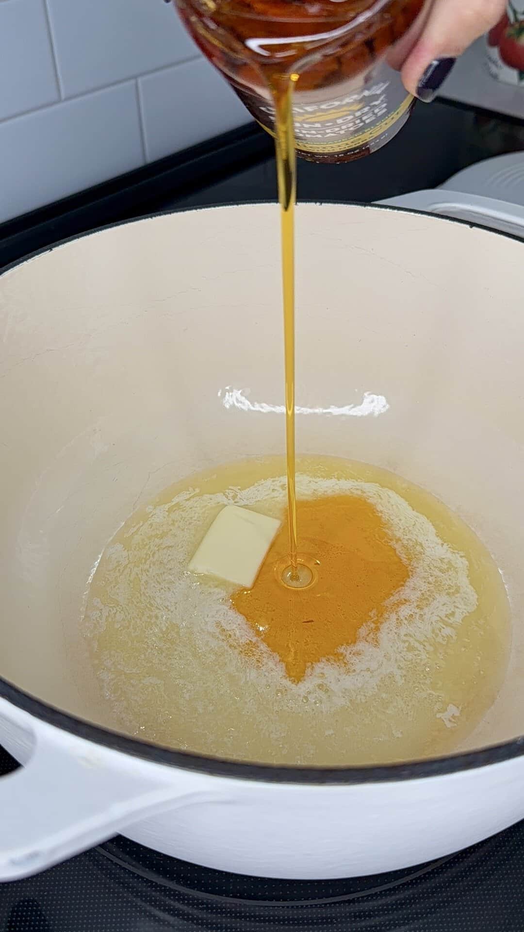A hand pours golden oil from a jar into a pot containing a melting pat of butter.