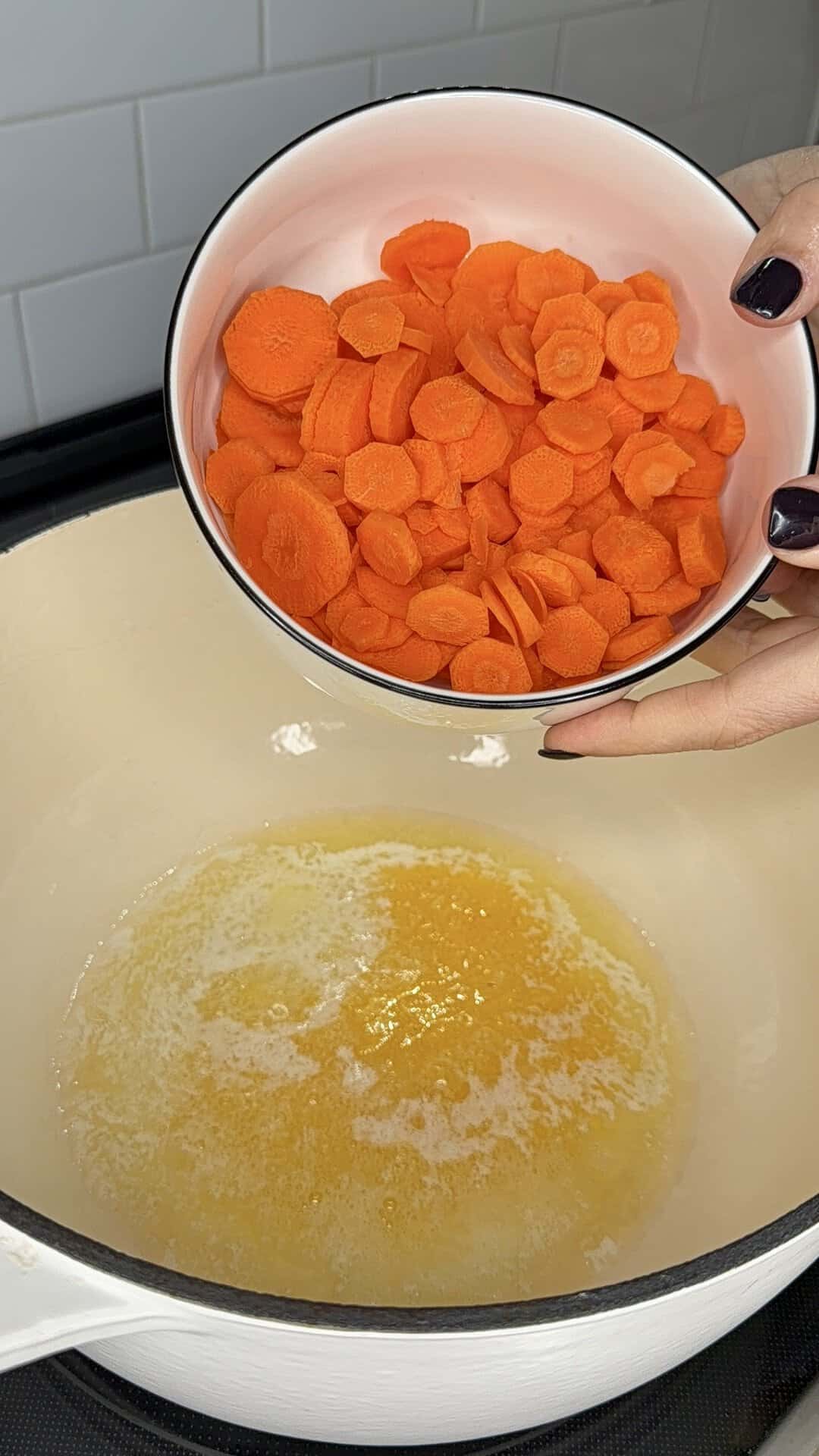 A hand pours a bowl of sliced carrots into a pot of melted butter.