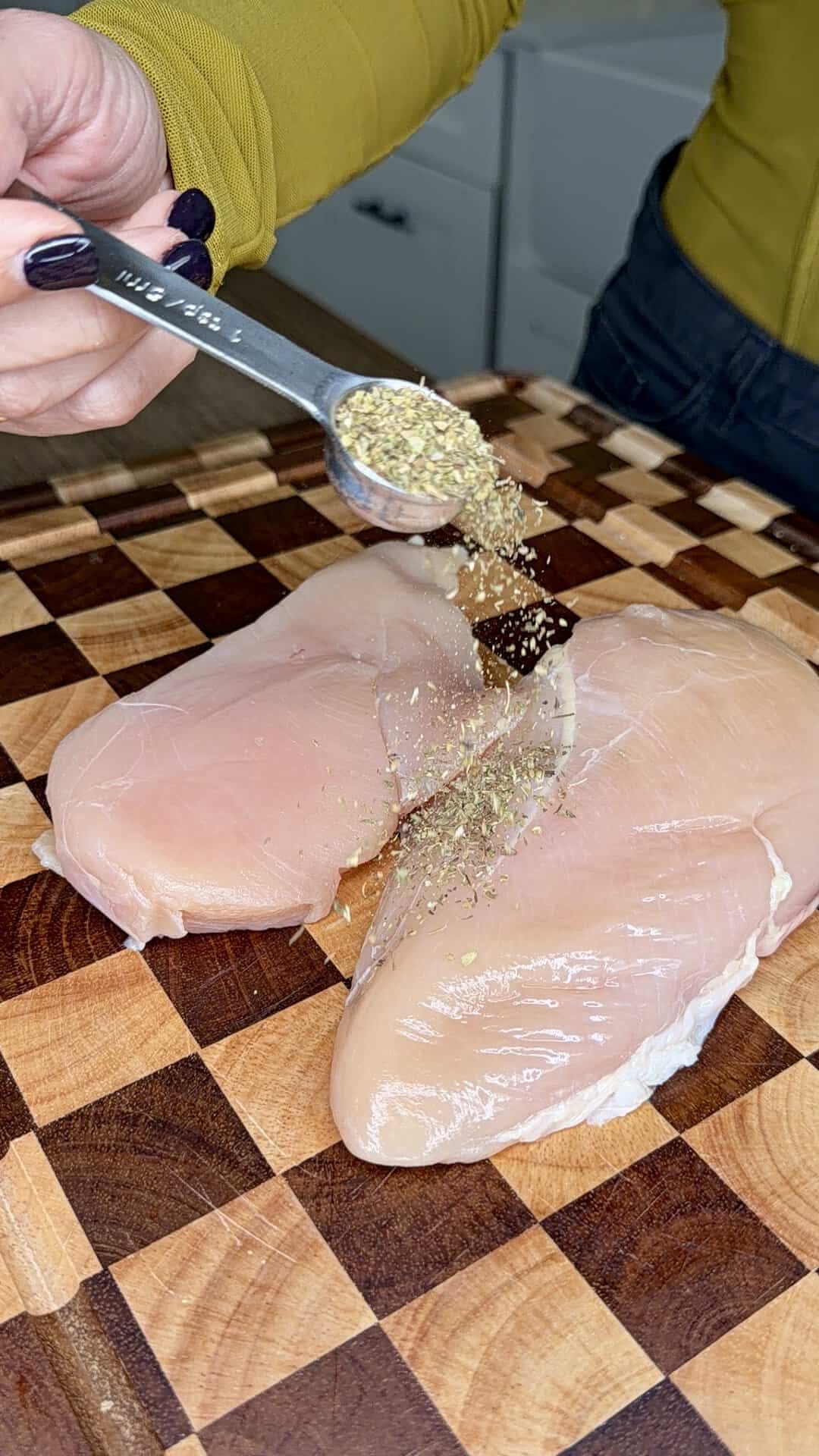 A person sprinkles dried herbs from a measuring spoon onto two raw chicken breasts on a wooden cutting board.
