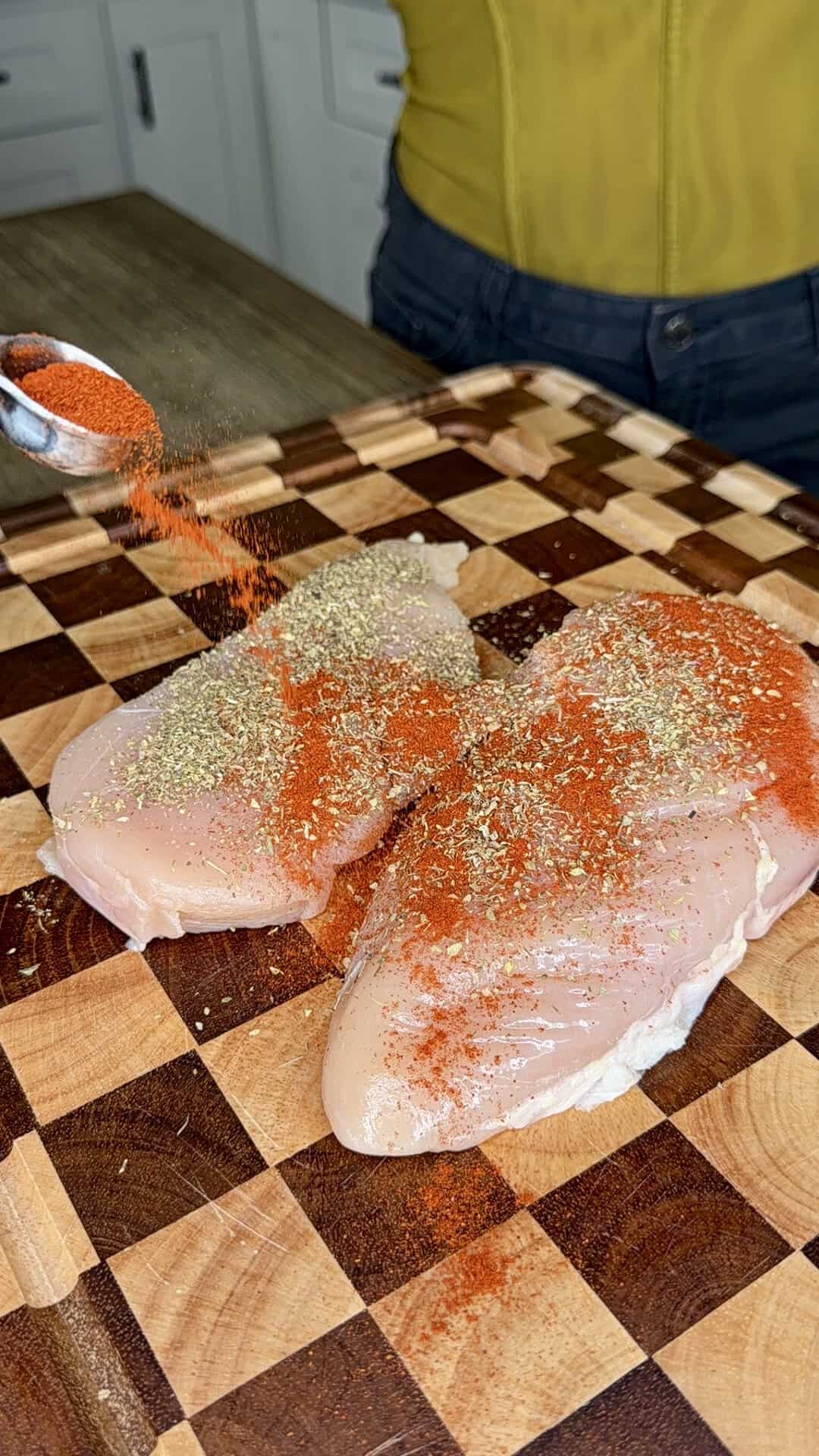 Red spices being sprinkled from a measuring spoon onto two raw chicken breasts on a wooden cutting board.