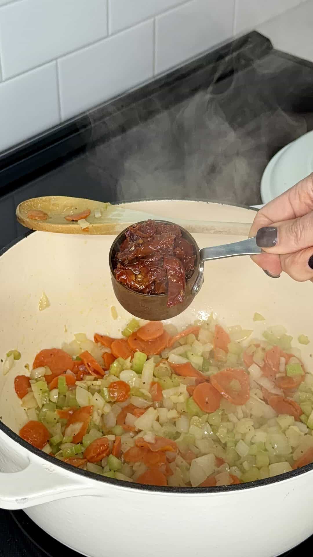 A hand holds a measuring cup filled with sun-dried tomatoes over a pot of steaming sautéed carrots, celery, and onions.
