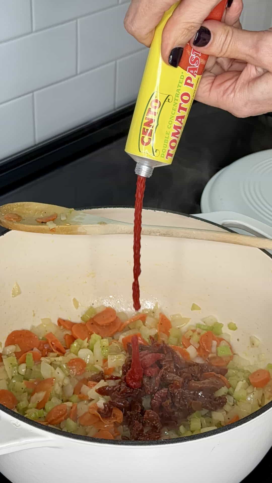 A hand squeezing tomato paste from a yellow Cento tube into a pot of sautéed carrots, celery, and onions.