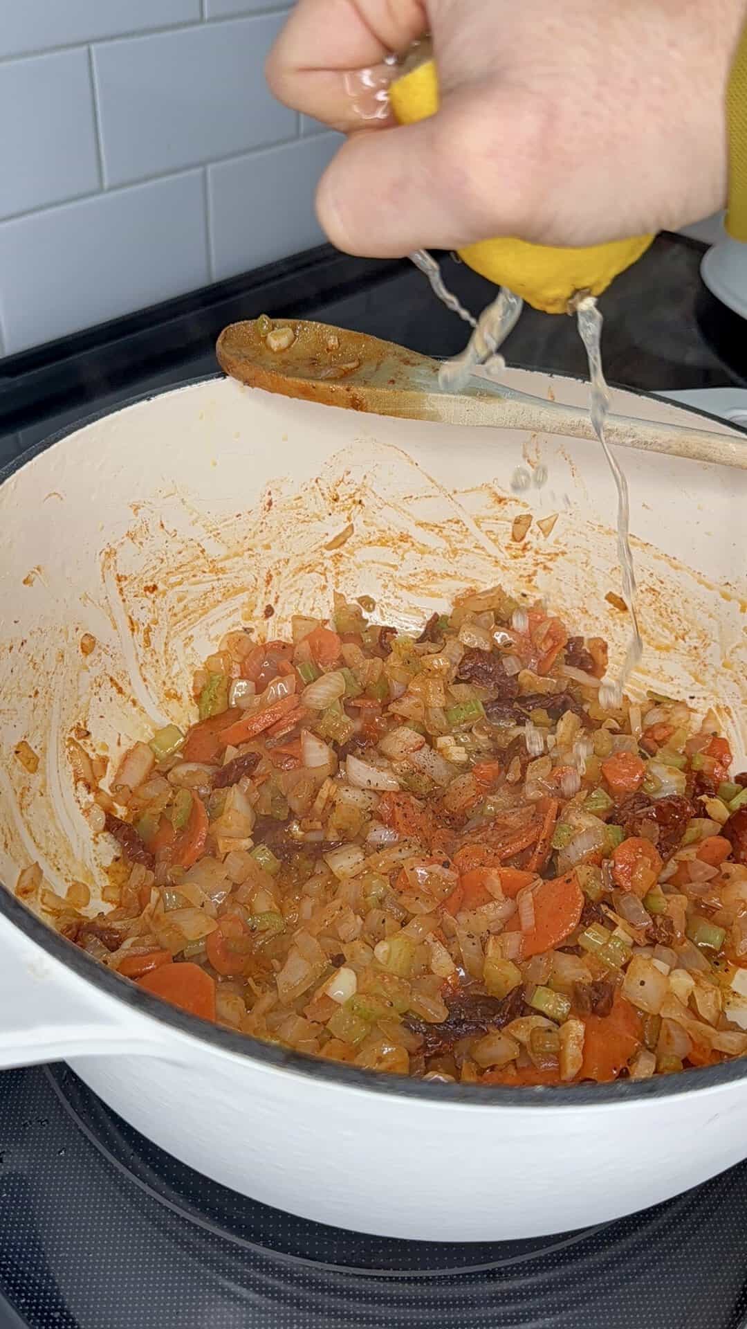 A hand squeezes a lemon over a Dutch oven filled with a mixture of sautéed carrots, celery, onions, and sun-dried tomatoes.