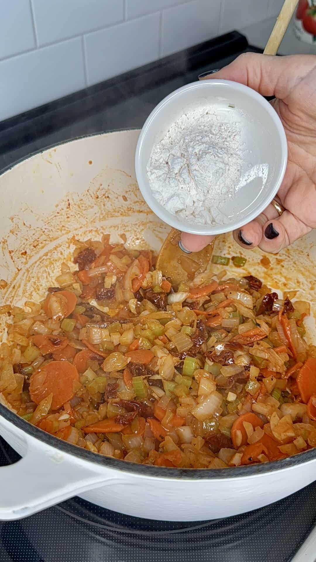A hand holds a small bowl of flour over a Dutch oven containing a sautéed mixture of carrots, celery, onions, and sun-dried tomatoes.