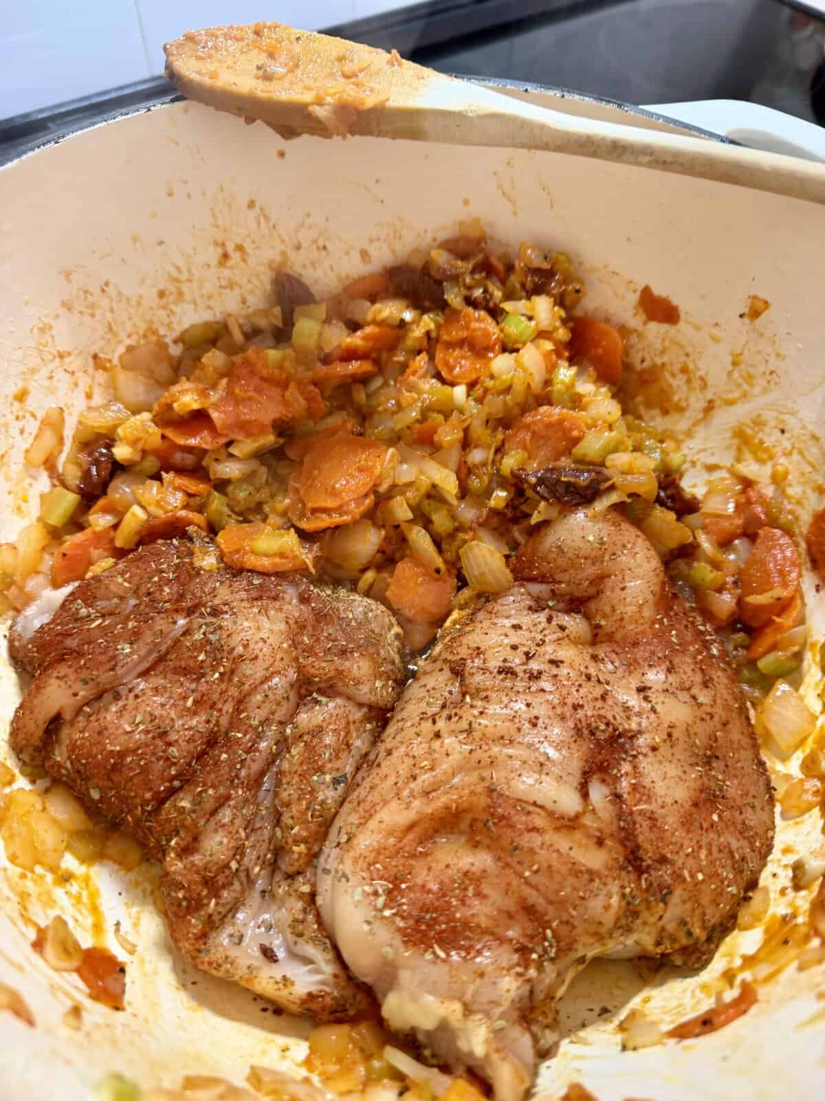 Two seasoned, raw chicken breasts resting in a Dutch oven with sautéed carrots, onions, and celery, with a wooden spoon resting on the rim.