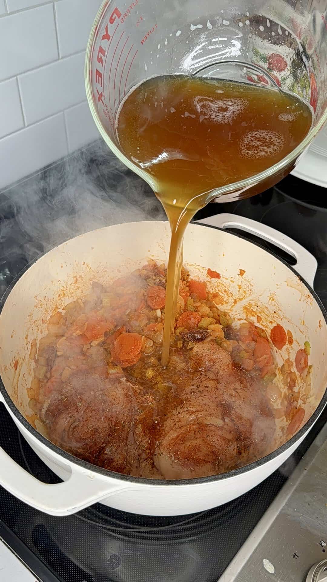 Broth being poured from a glass measuring cup into a white pot containing seared meat and sautéed vegetables.