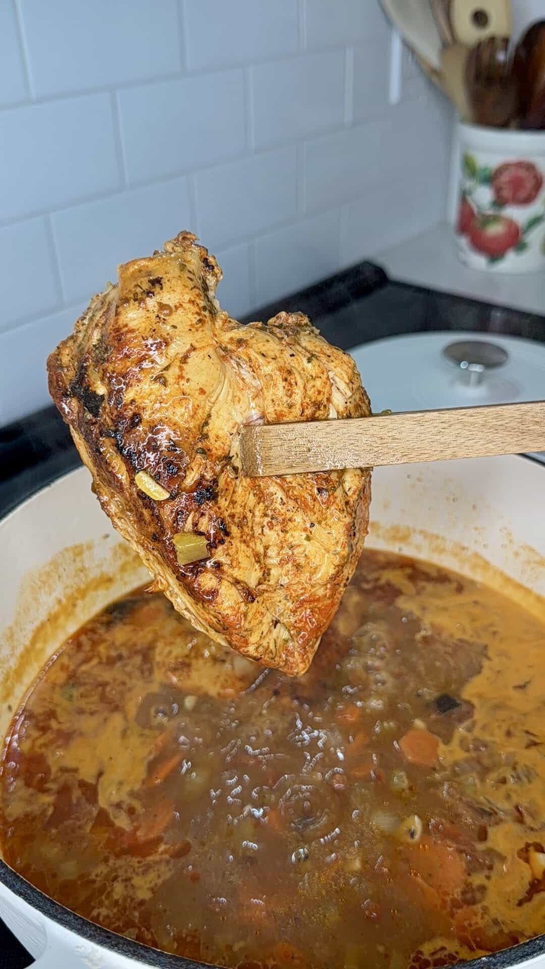 Wooden tongs hold a seasoned, seared chicken breast over a pot of simmering vegetable stew.