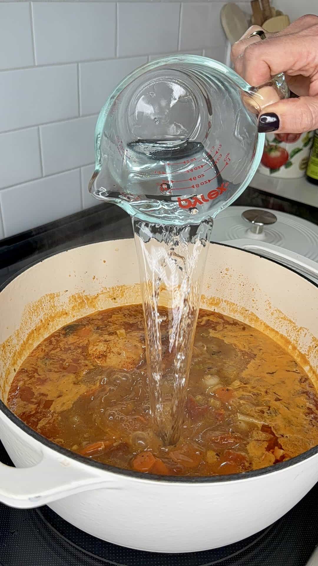 Water being poured from a glass measuring cup into a white Dutch oven filled with a red vegetable soup.
