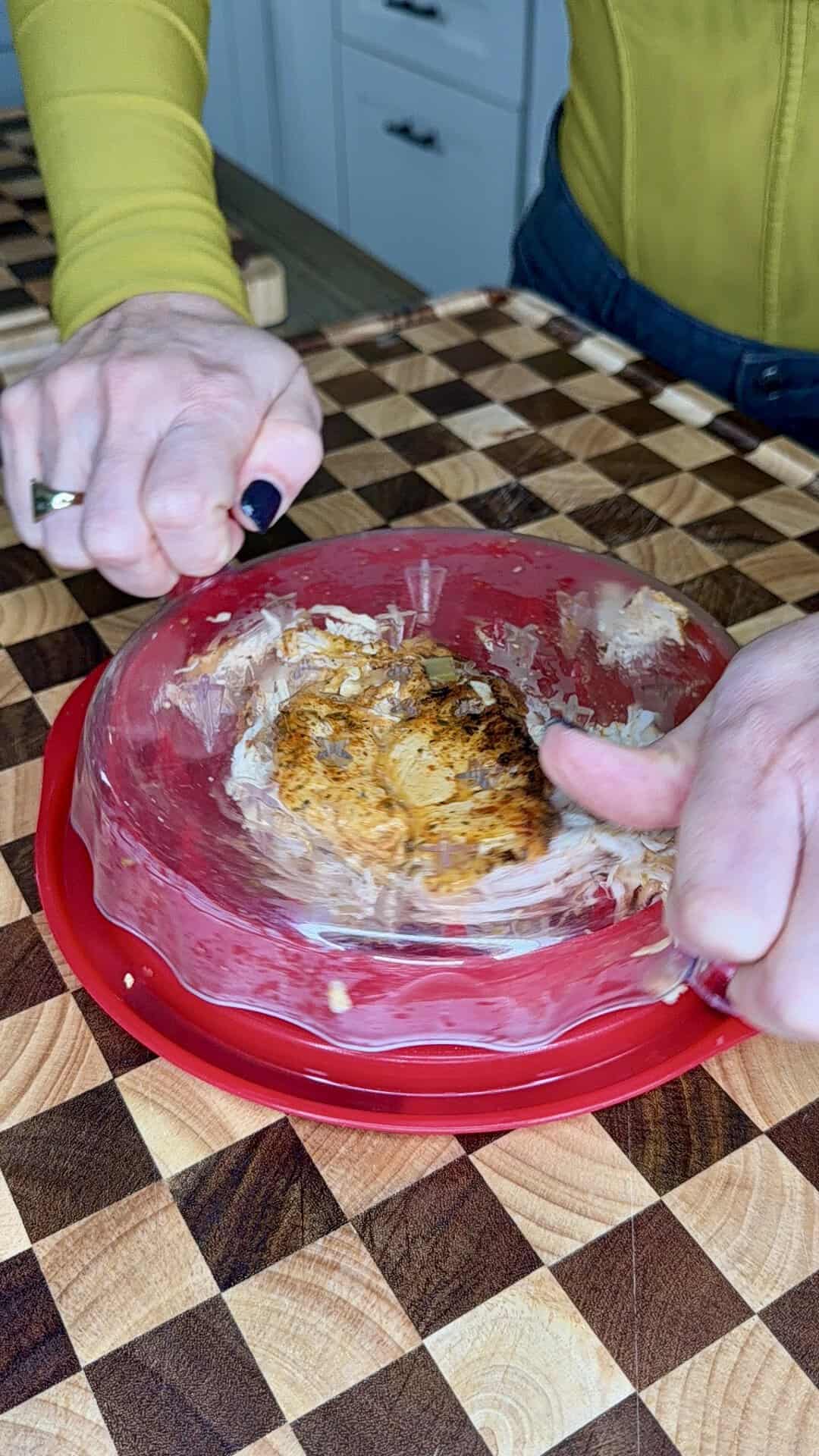 A person presses a clear plastic lid onto a cooked chicken breast on a checkered cutting board.