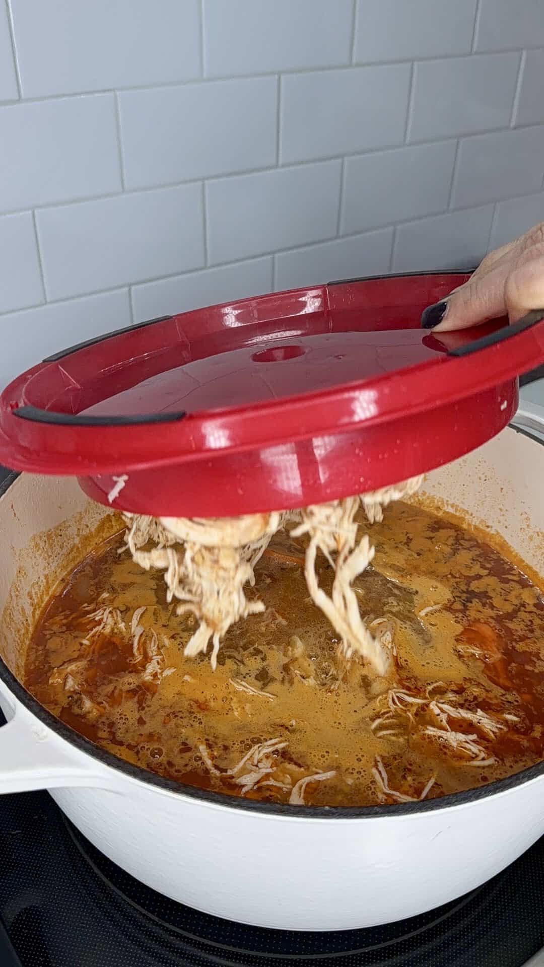 A hand holds a red lid over a pot of stew, with shredded chicken hanging from the underside of the lid into the broth.
