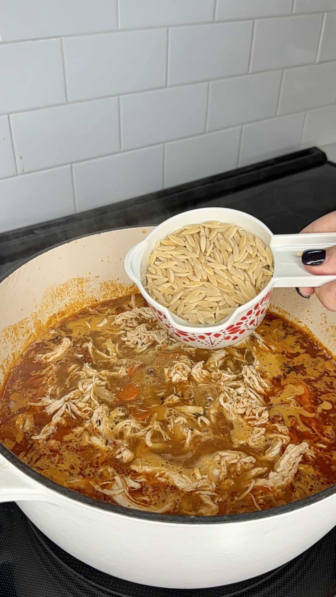 A hand holds a measuring cup filled with dry orzo over a large pot of simmering chicken soup.