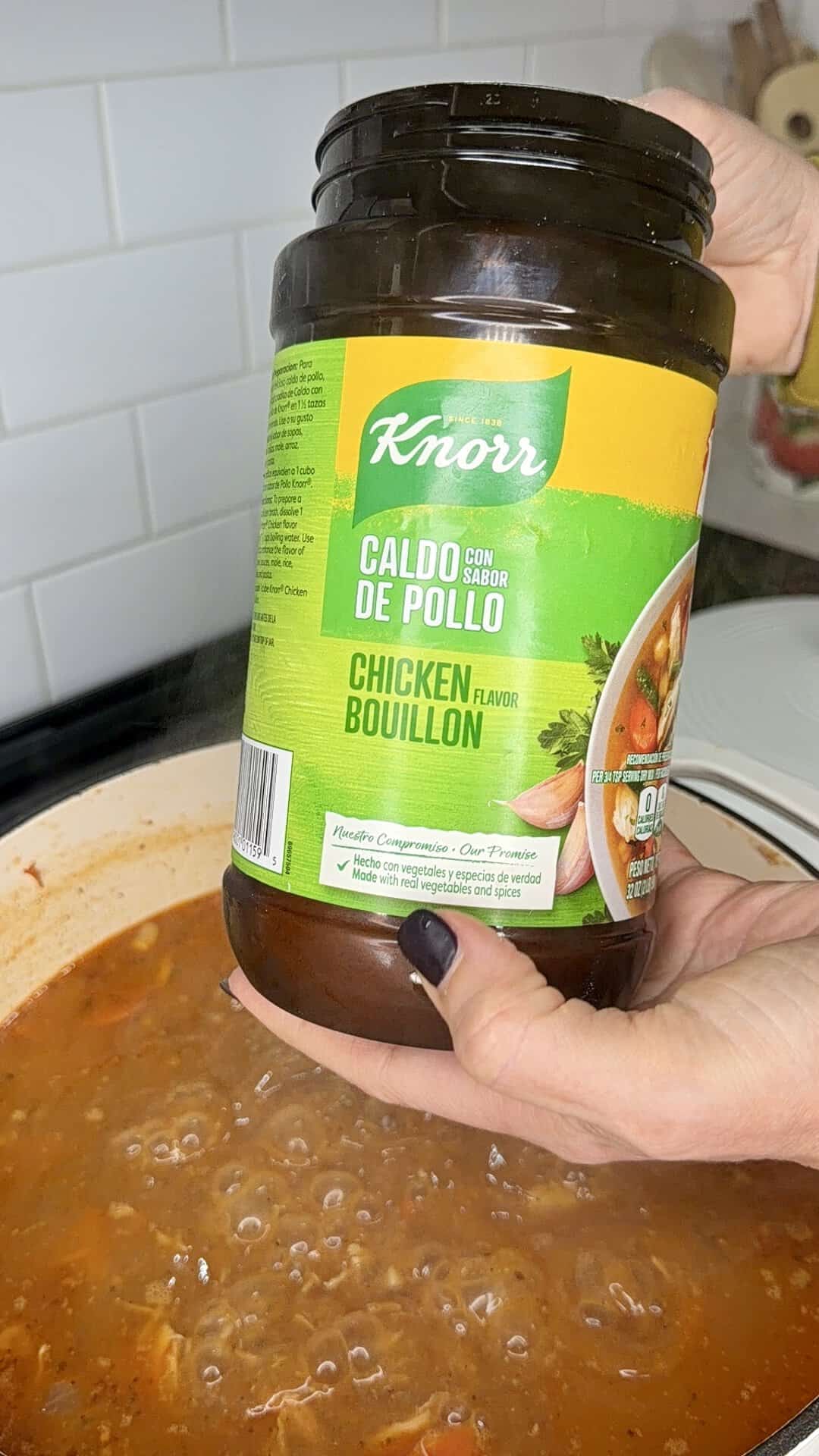 A hand holding a jar of Knorr chicken flavor bouillon over a pot of simmering soup.