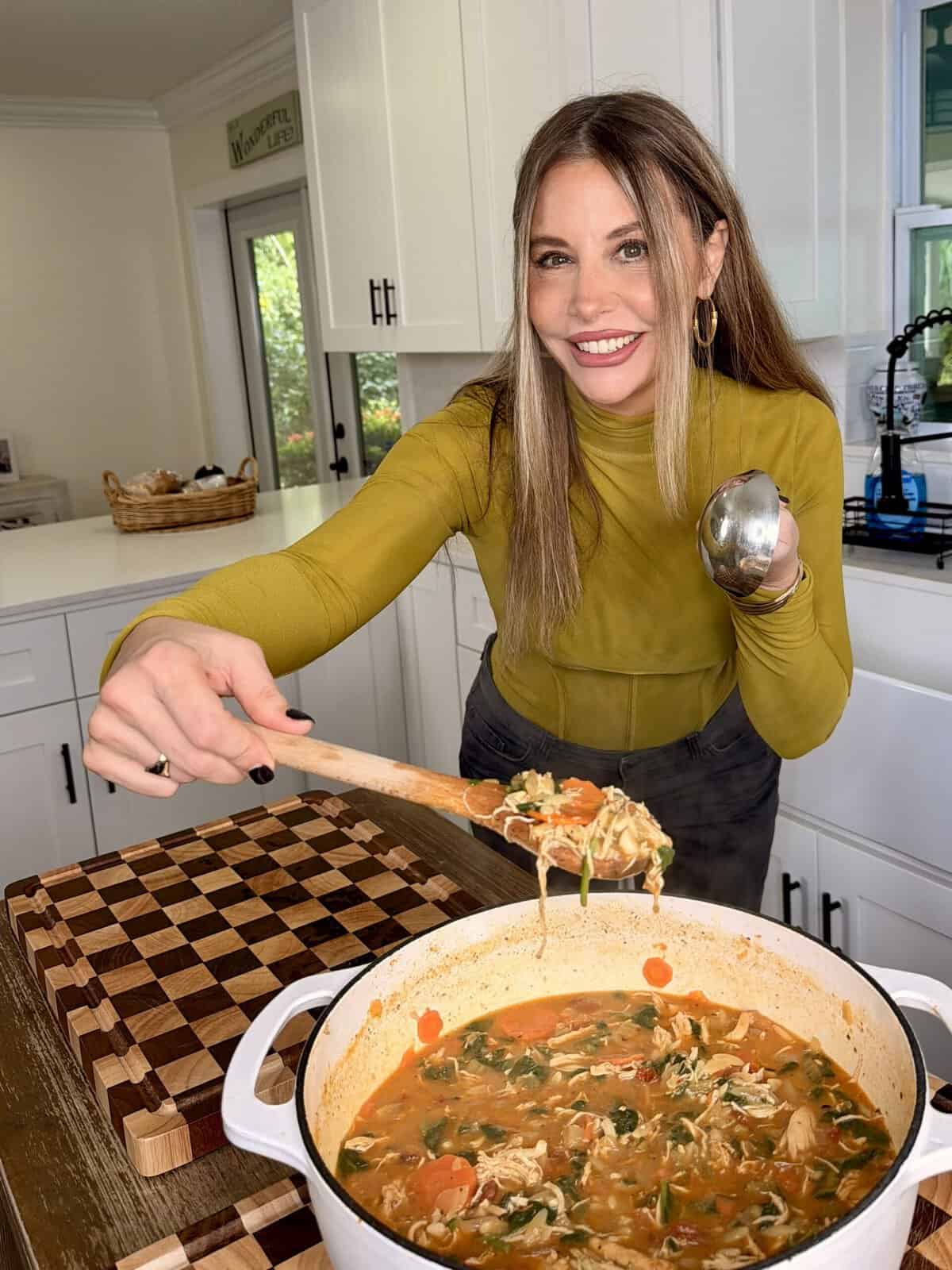 A woman in a mustard yellow top holds a wooden spoon with a scoop of chicken soup over a large pot of the same soup.