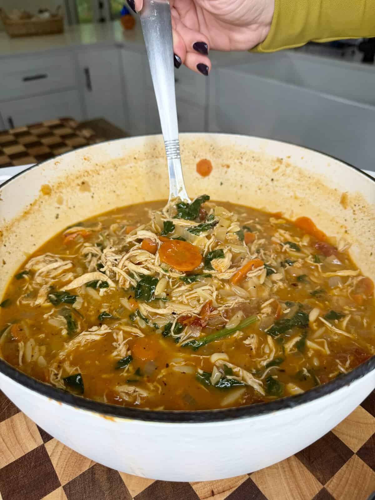 A hand holds a spoonful of chicken and vegetable soup in a white Dutch oven.