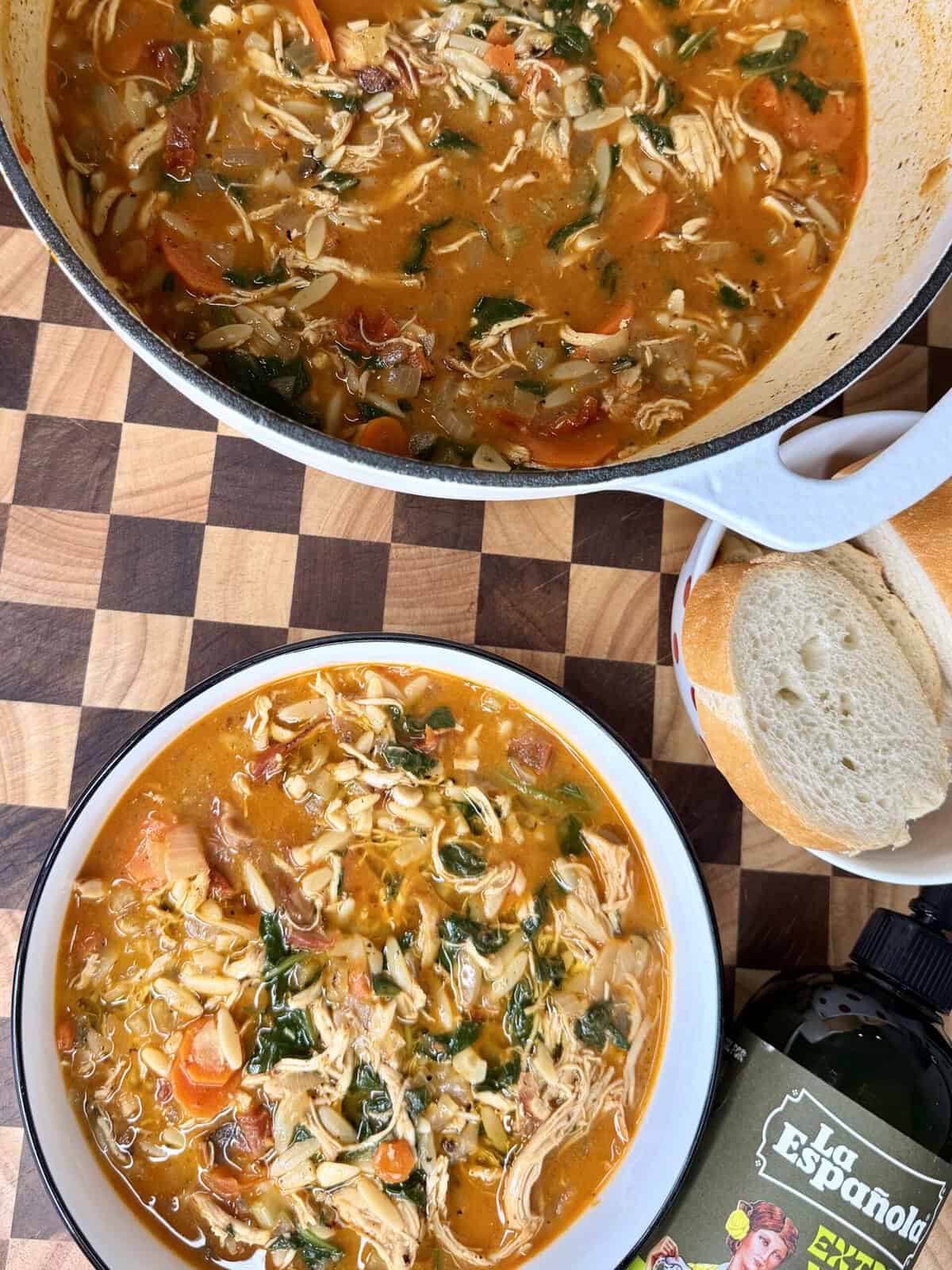 A bowl of chicken and vegetable soup with orzo next to a pot of the same soup, a side of bread, and a bottle of olive oil on a checkered cutting board.