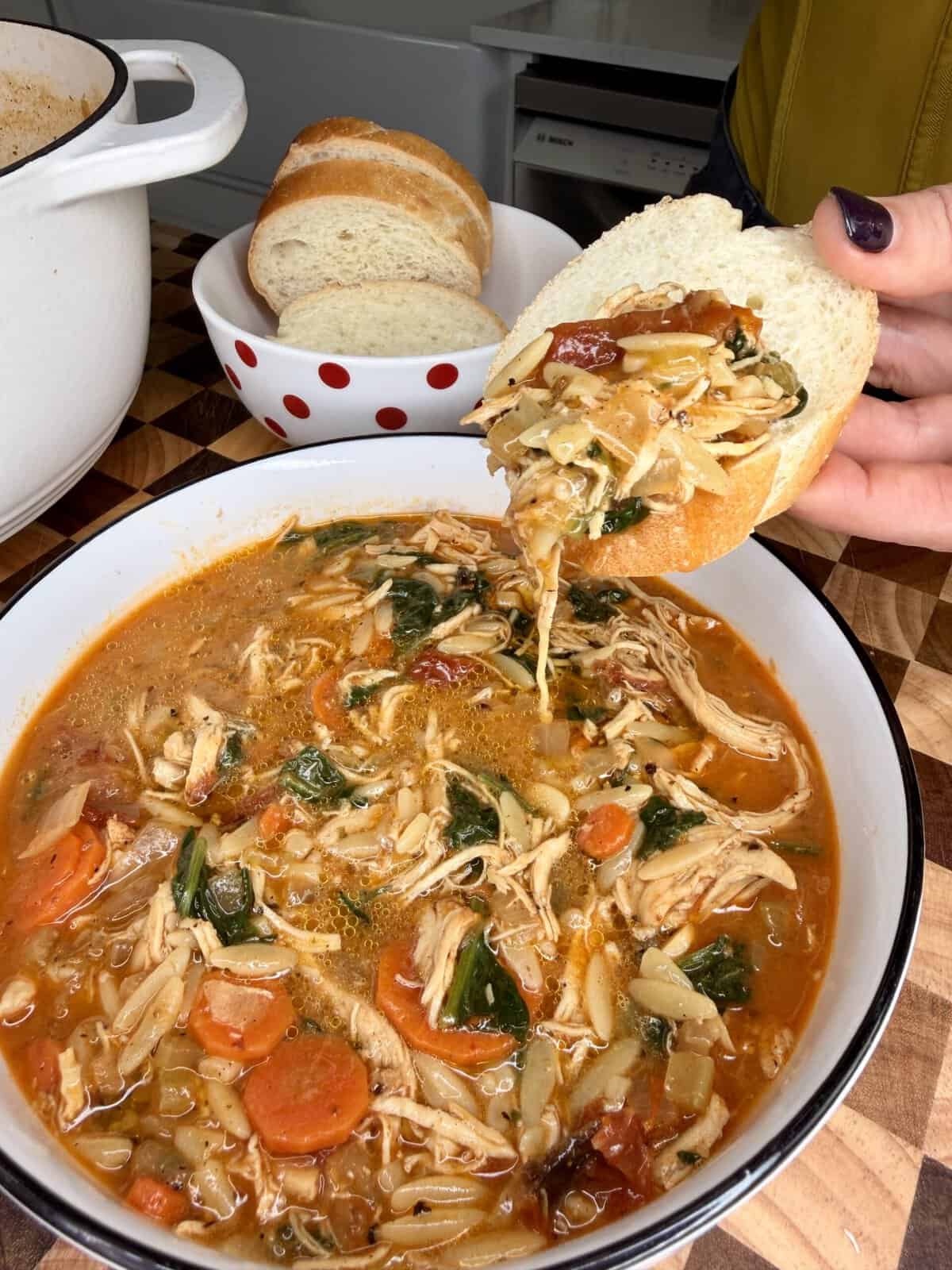 A hand dips a slice of bread into a bowl of chicken, orzo, and vegetable soup.