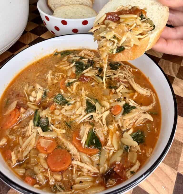 A hand dips a slice of crusty bread into a bowl of chicken orzo soup with carrots and greens, with a small bowl of sliced bread in the background.