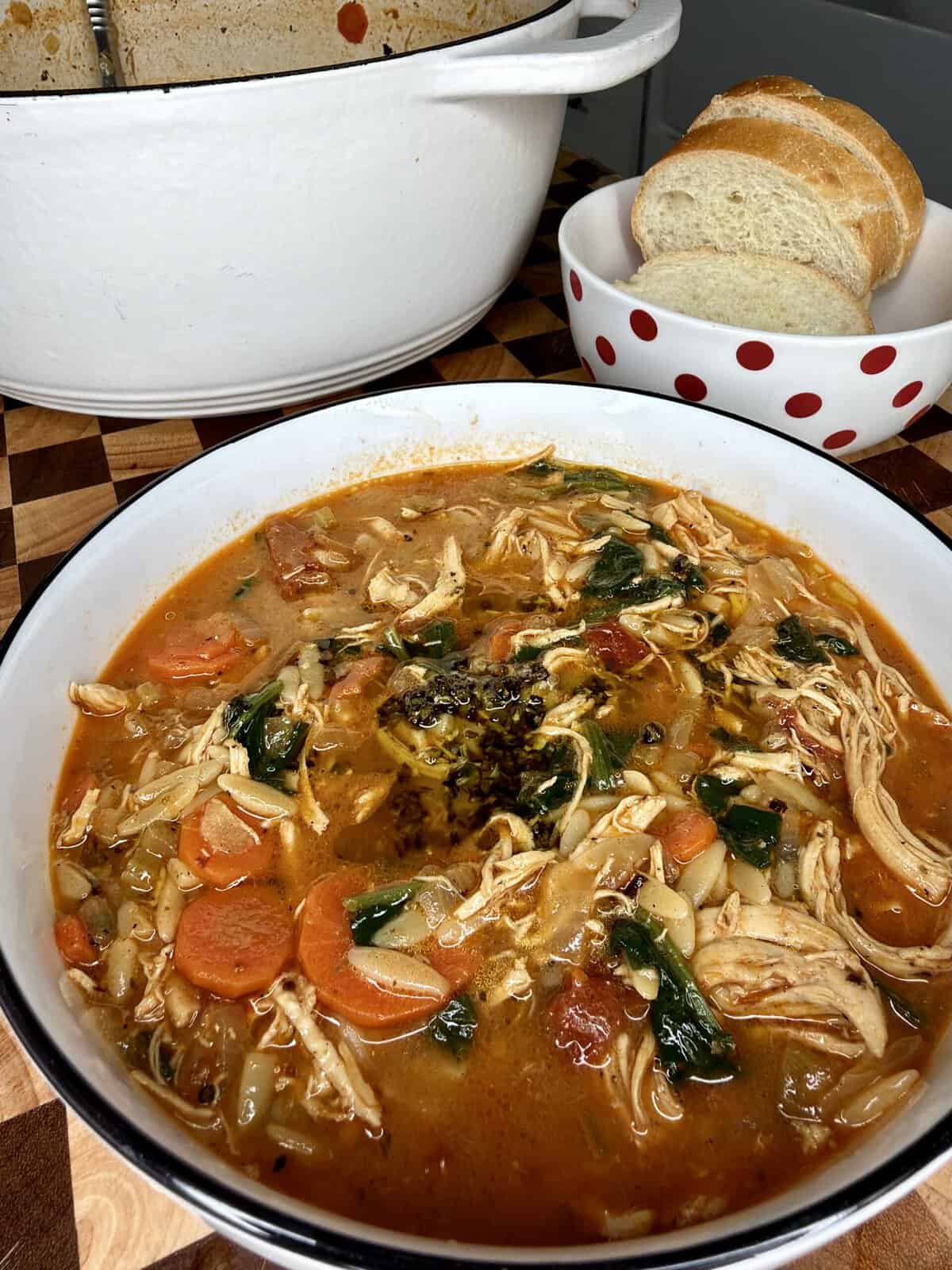 A bowl of chicken and vegetable soup with orzo and herbs, served with slices of bread in a small polka-dot bowl.