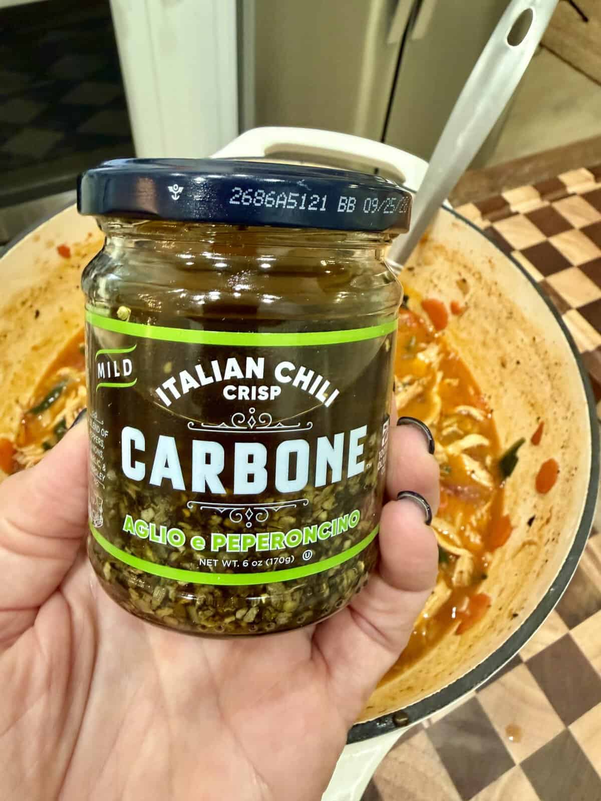 A hand holding a jar of Carbone Italian Chili Crisp Aglio e Peperoncino over a pot of cooked pasta.
