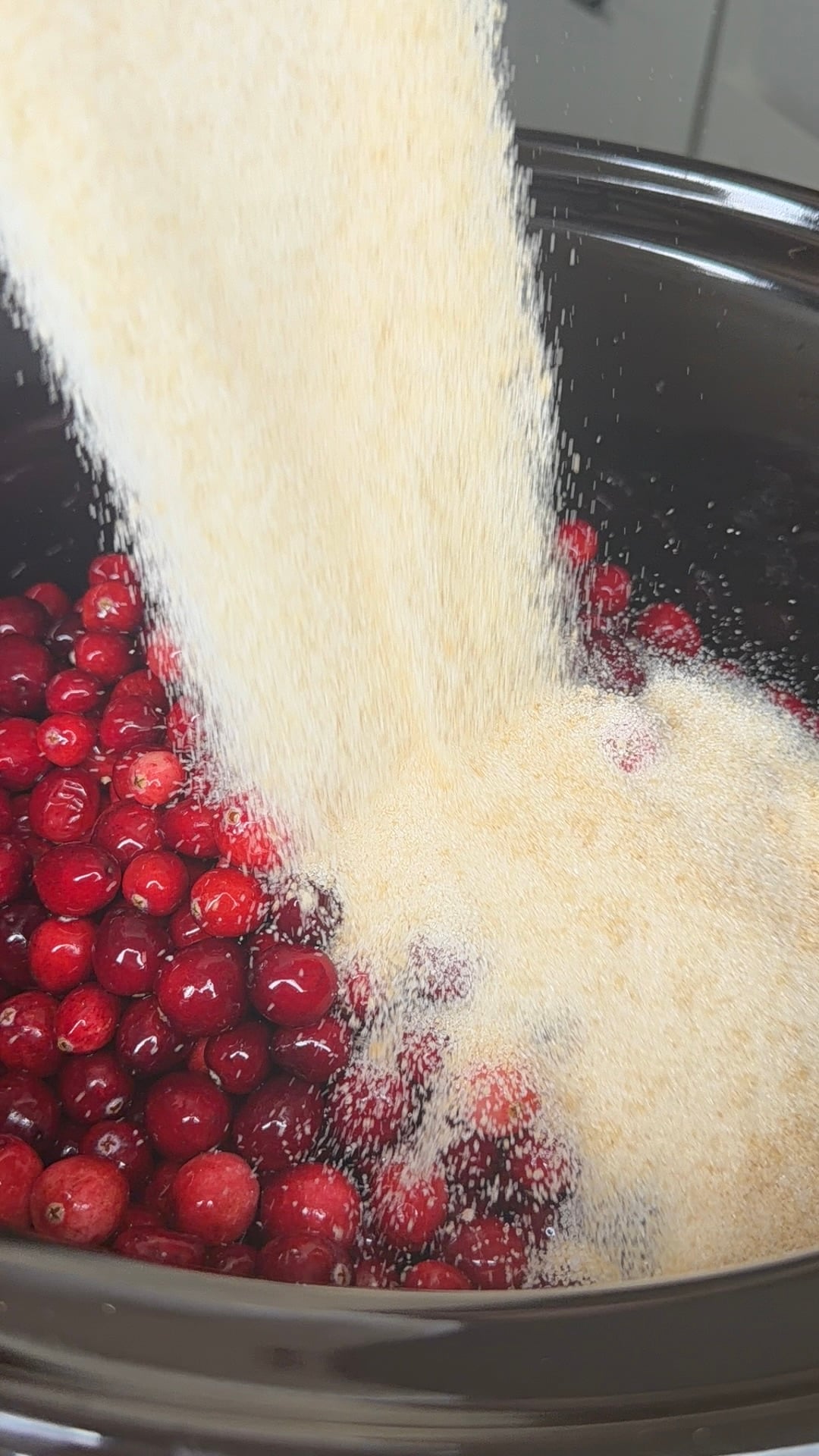 Granulated sugar being poured into a slow cooker filled with fresh cranberries.