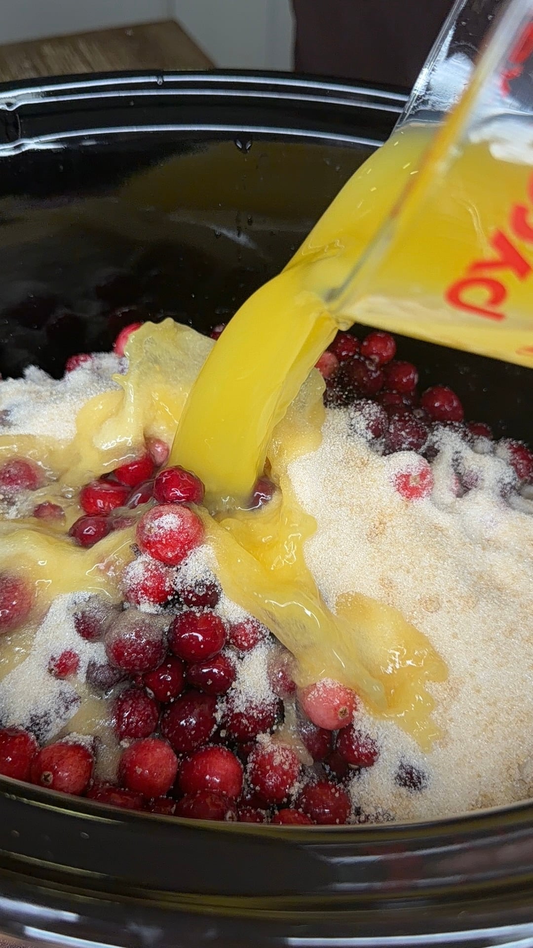 Orange juice being poured from a measuring cup into a slow cooker filled with fresh cranberries and granulated sugar.