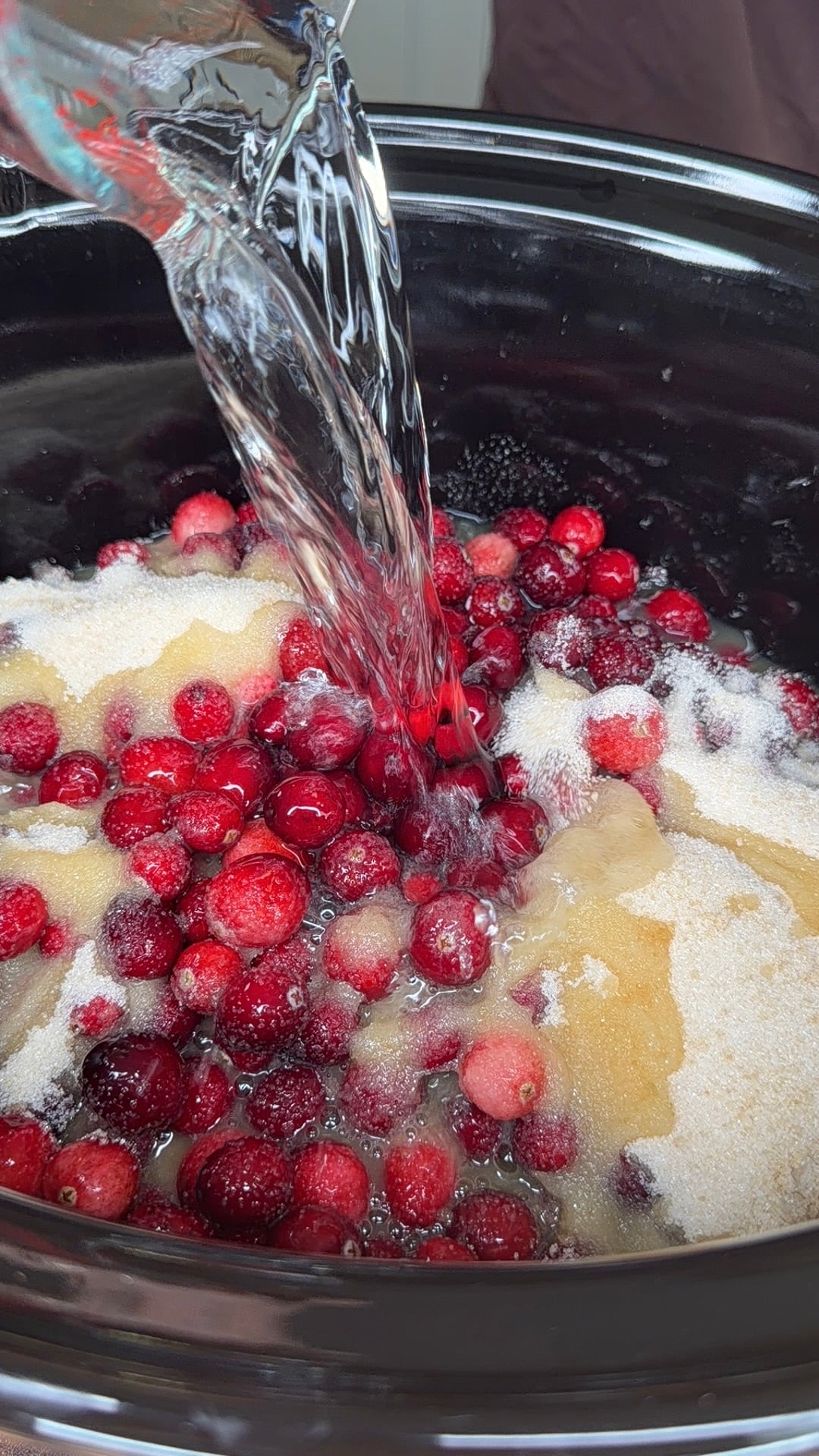 Water being poured into a slow cooker filled with fresh cranberries and sugar.