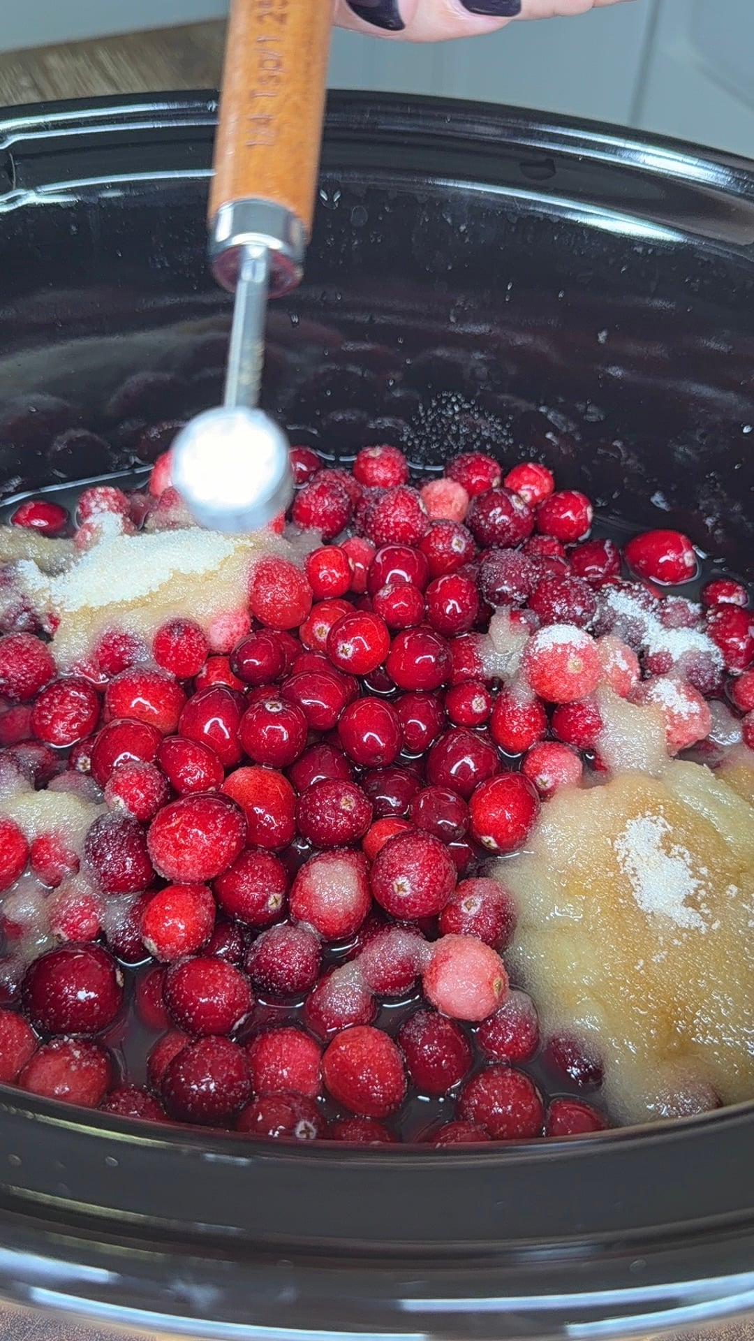 A hand holds a measuring spoon over a slow cooker filled with fresh cranberries and sugar.