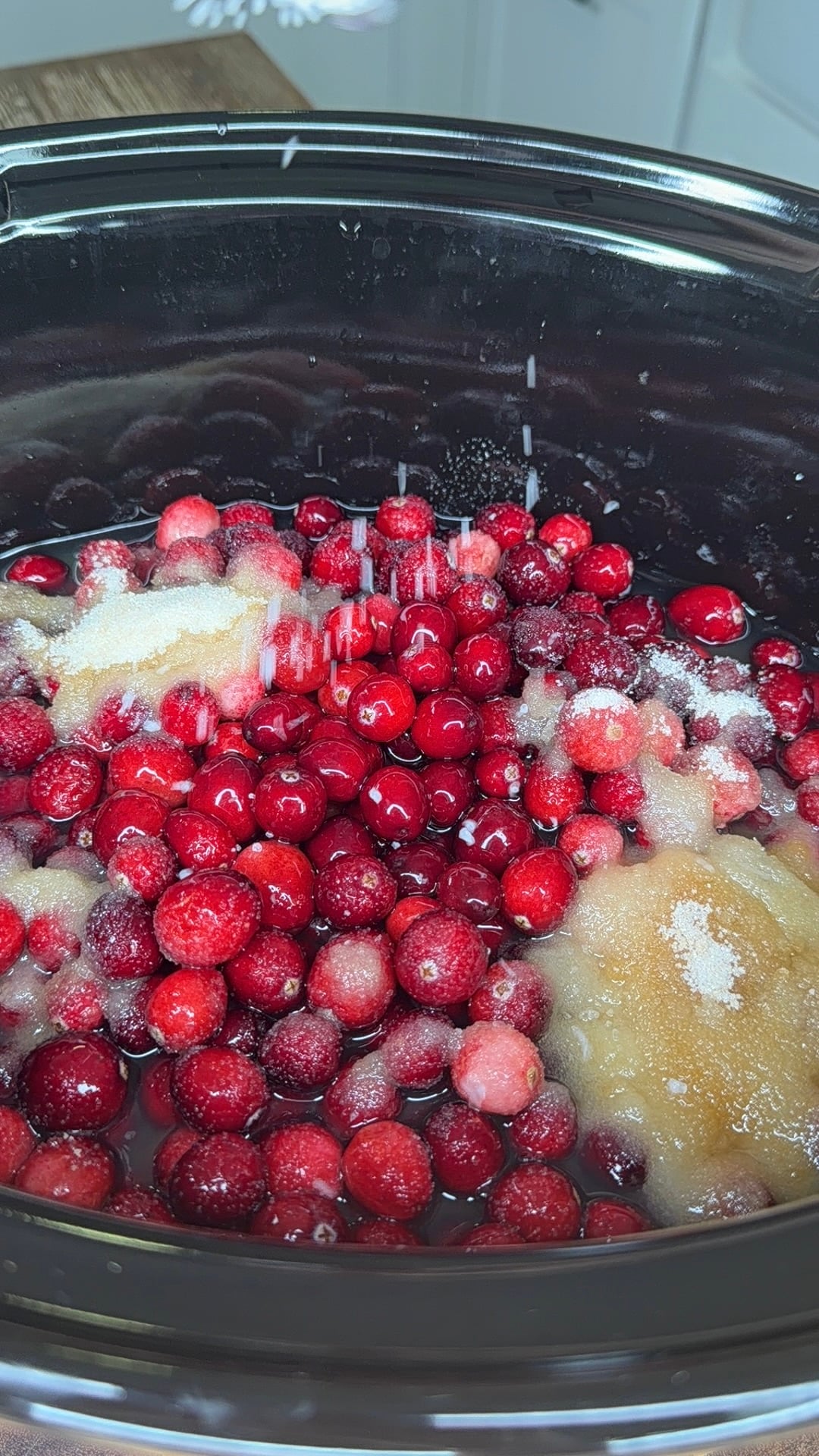 Fresh cranberries and chunks of fruit in a slow cooker, with sugar being sprinkled over the top.