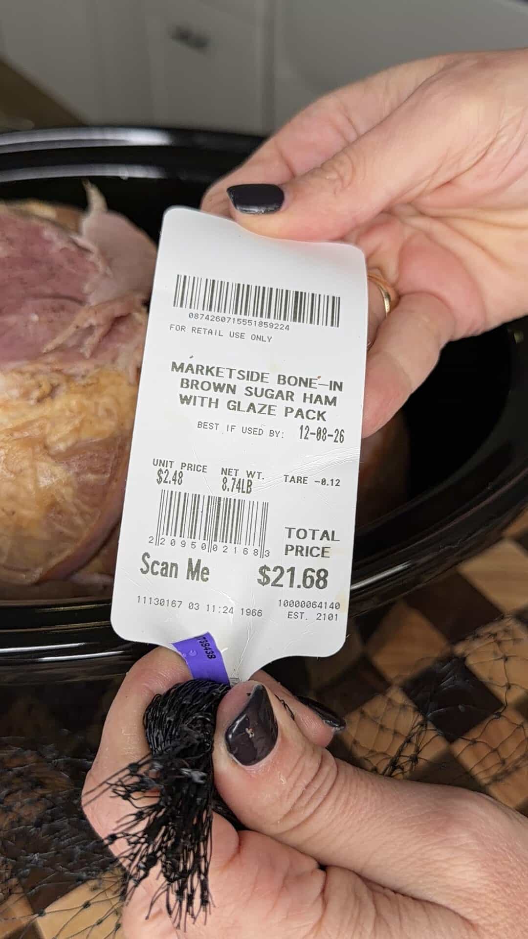 Hands hold a grocery store label for a Marketside bone-in brown sugar ham with a total price of $21.68, positioned in front of a ham in a slow cooker.