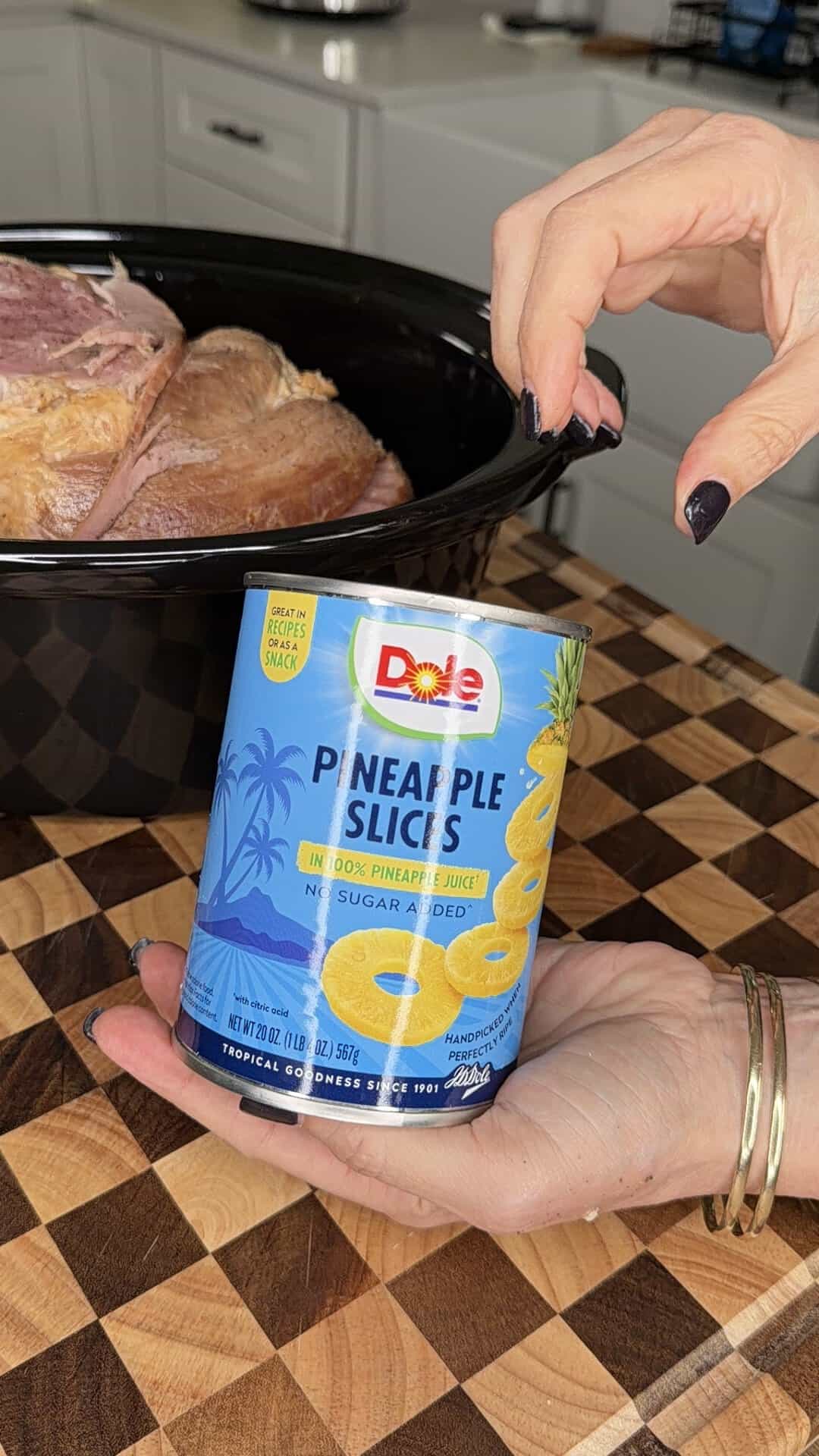 A hand holds a can of Dole pineapple slices in front of a slow cooker containing a cooked ham.