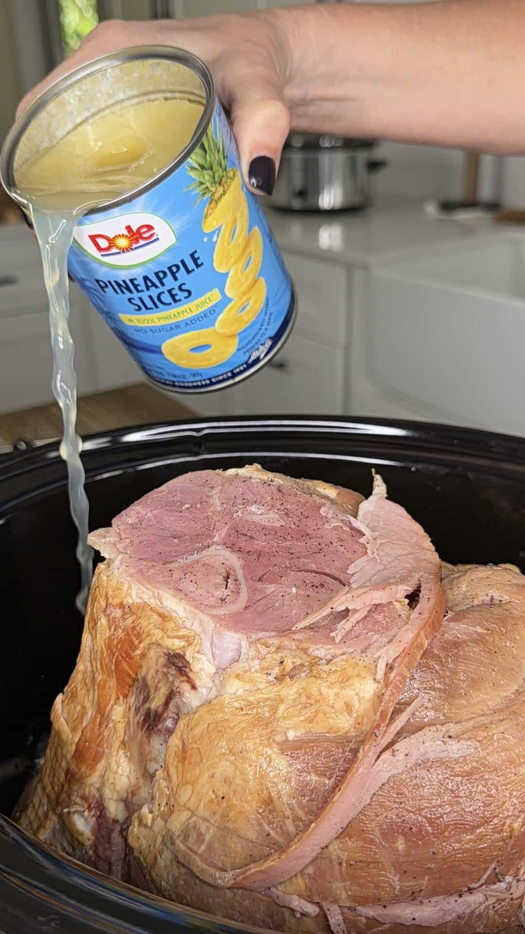 A hand pours pineapple juice from a Dole pineapple slices can over a large ham in a slow cooker.