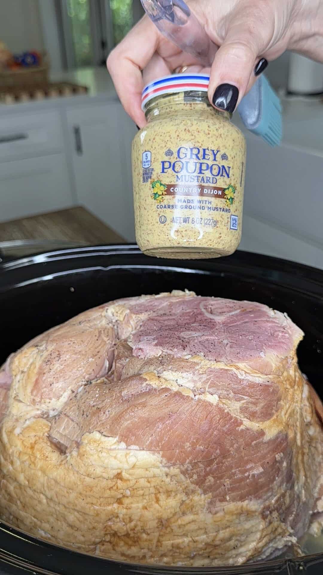 A hand holds a jar of Grey Poupon Country Dijon mustard over a large ham in a slow cooker.