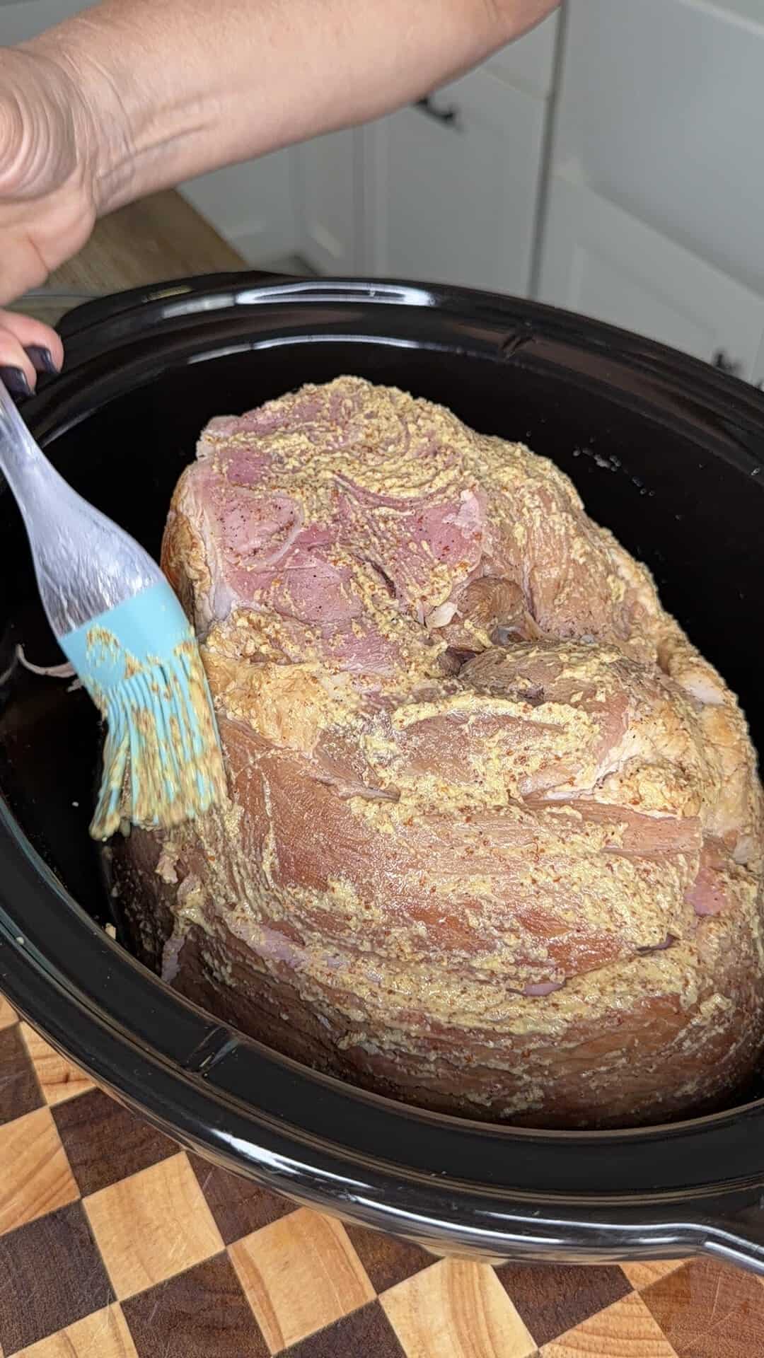 A hand uses a silicone brush to spread mustard glaze onto a large ham inside a slow cooker.