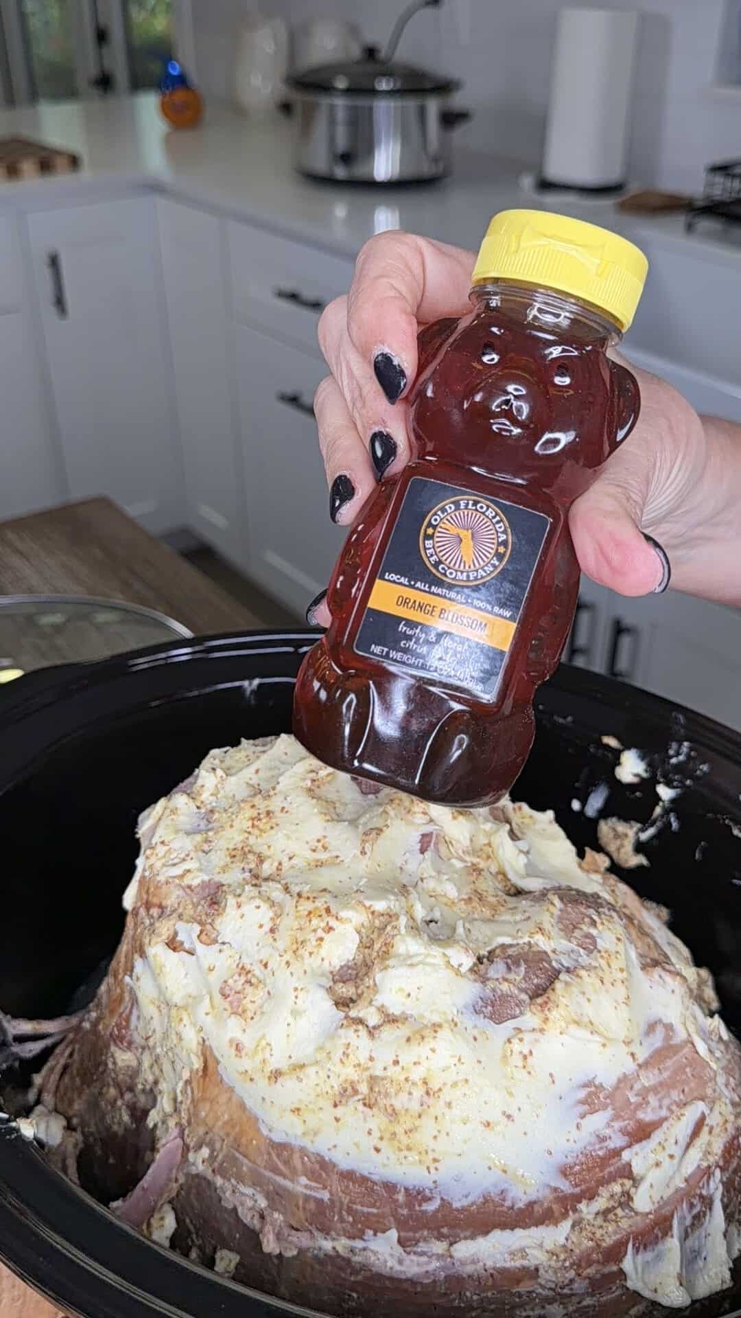 A hand pours Old Florida Company orange blossom honey from a bear-shaped bottle onto a raw ham covered in a white glaze inside a slow cooker.