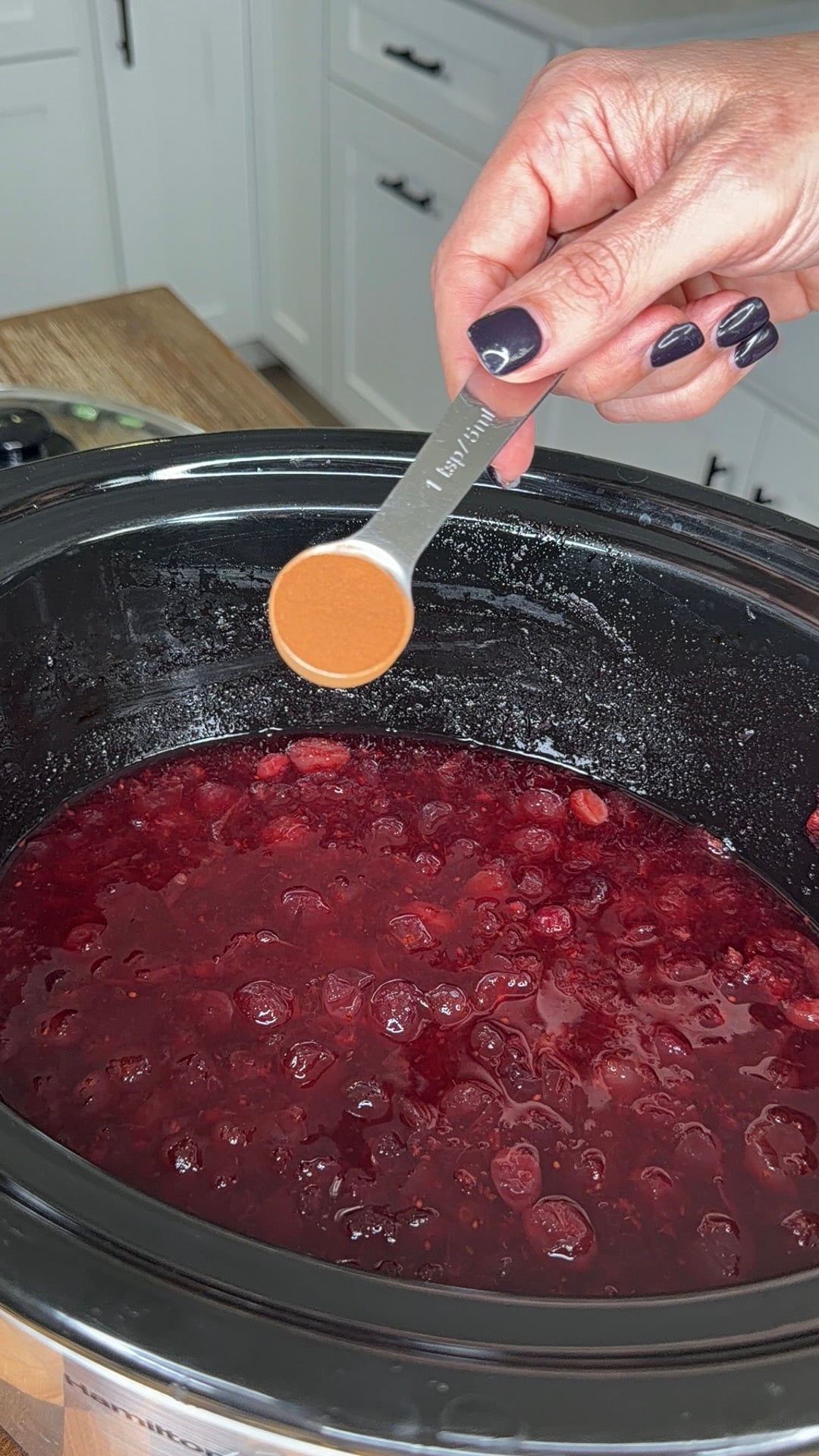 A hand holds a measuring spoon filled with cinnamon over a slow cooker filled with cranberry sauce.